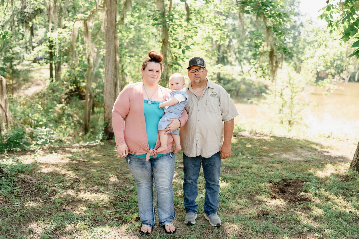 Family of three posing together for a portrait in a wooded setting on a sunny day.