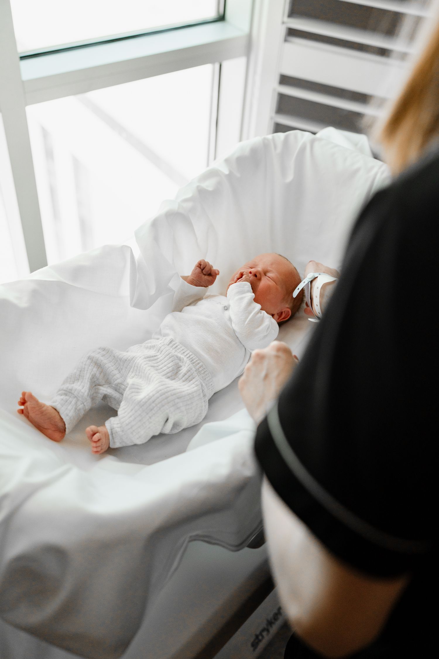 Caregiver's hands carefully positioning sleeping infant on white surface near window.