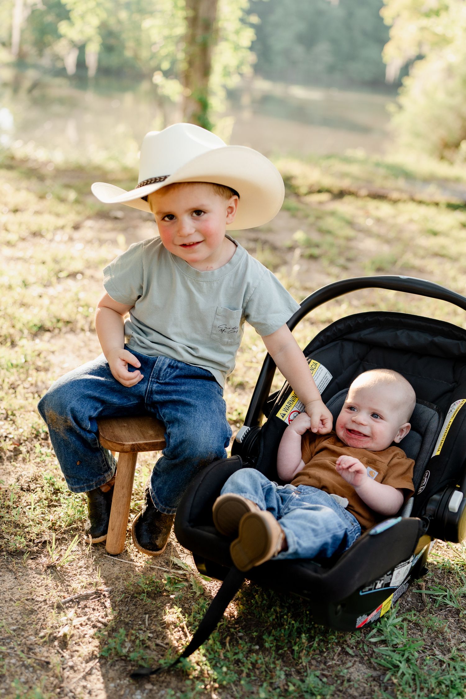 Two children interact playfully near a car seat in a wooded setting.