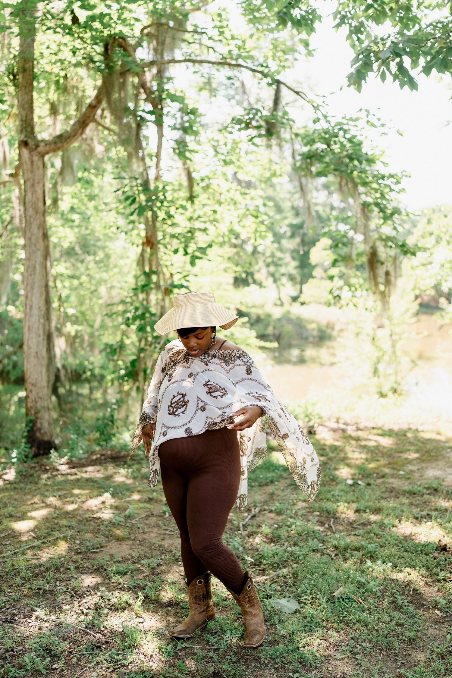 Woman in a wide-brimmed hat and patterned white poncho poses gracefully in a sunlit wooded area.