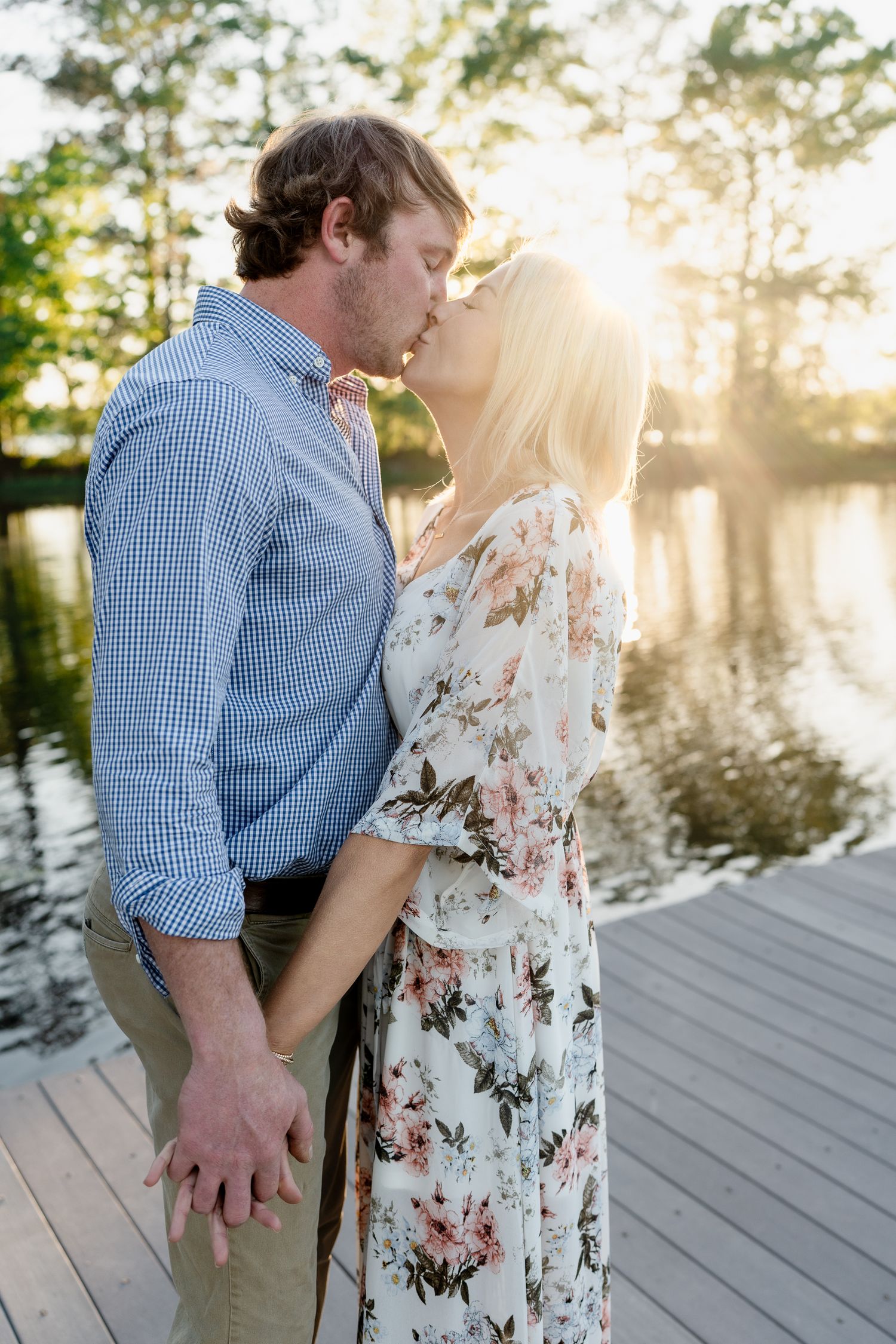 Couple embracing in a tender kiss on wooden dock with sunlit water and trees in background.