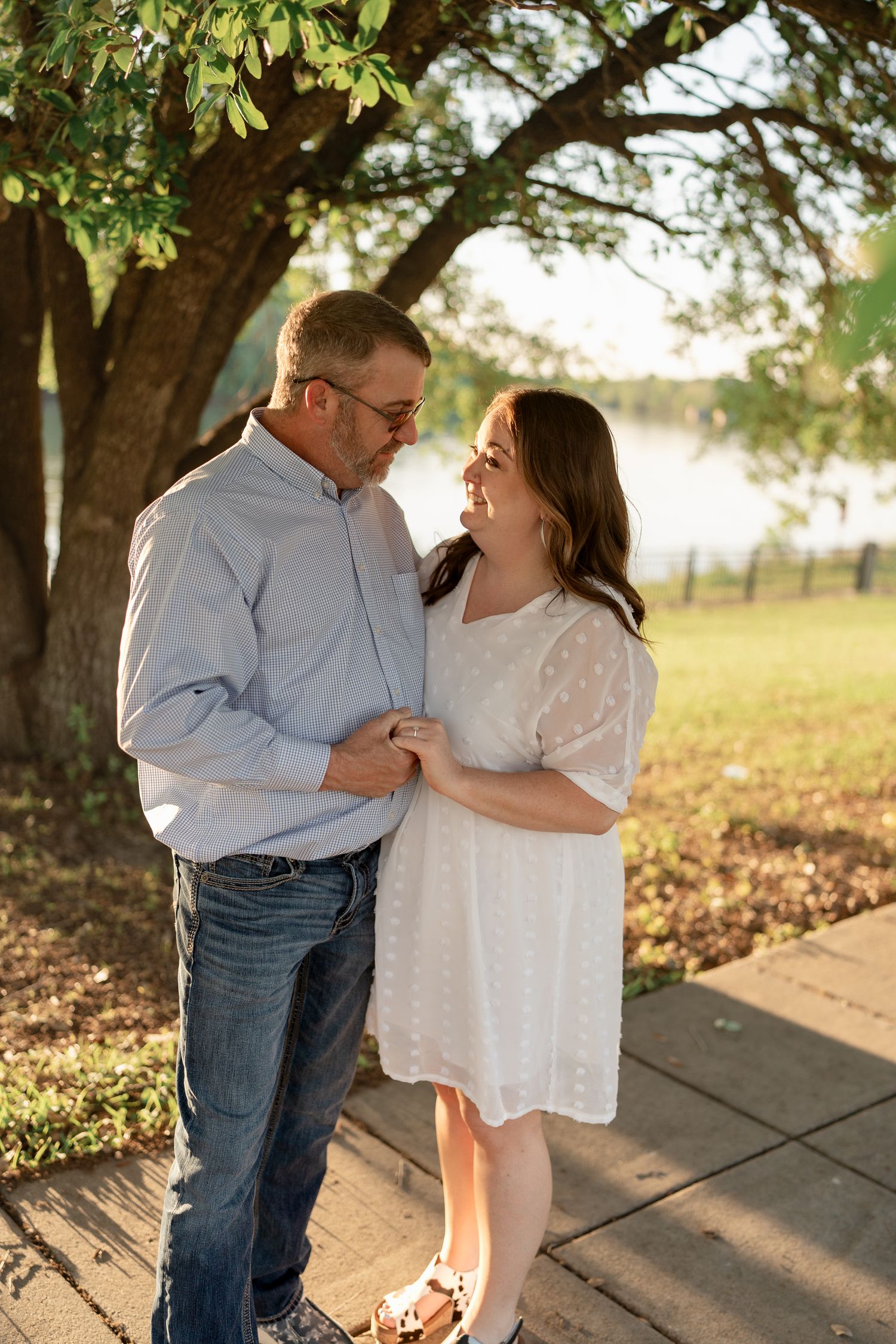 Couple sharing tender moment under tree canopy with sunlit water in background.