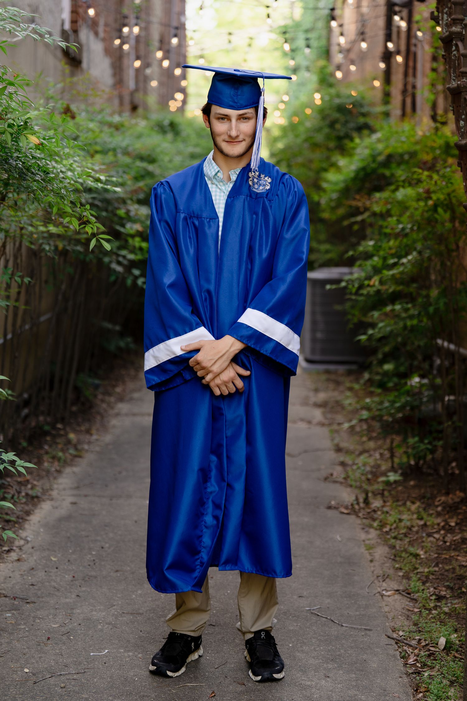 Graduate in full blue academic regalia stands on outdoor path with string lights.