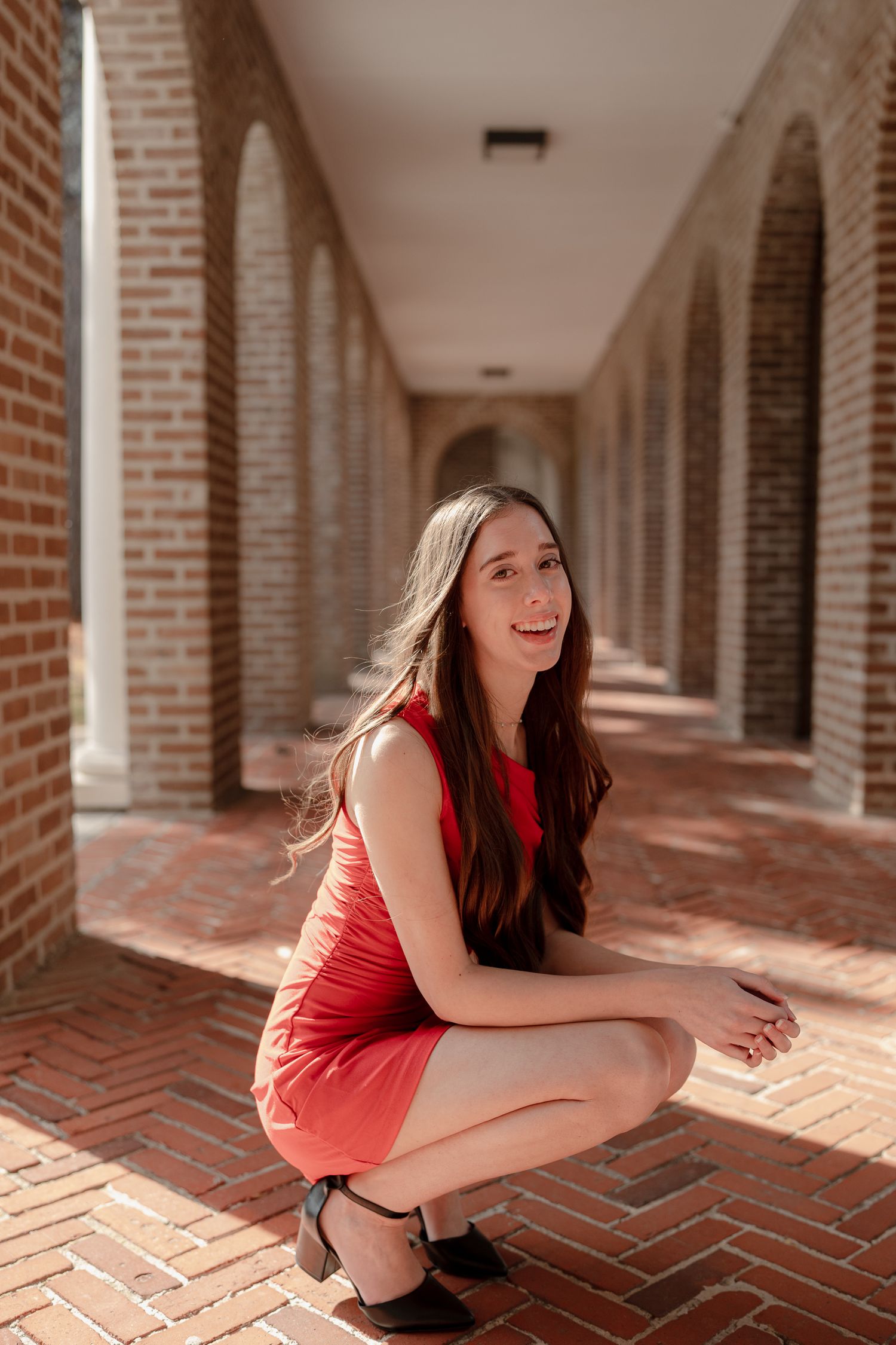 A person in a coral dress poses playfully in a sunlit brick archway corridor.