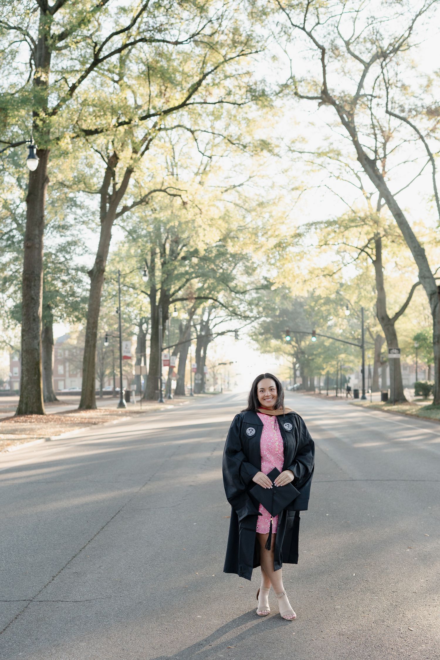 Graduate in academic regalia stands on tree-lined campus street during golden hour.