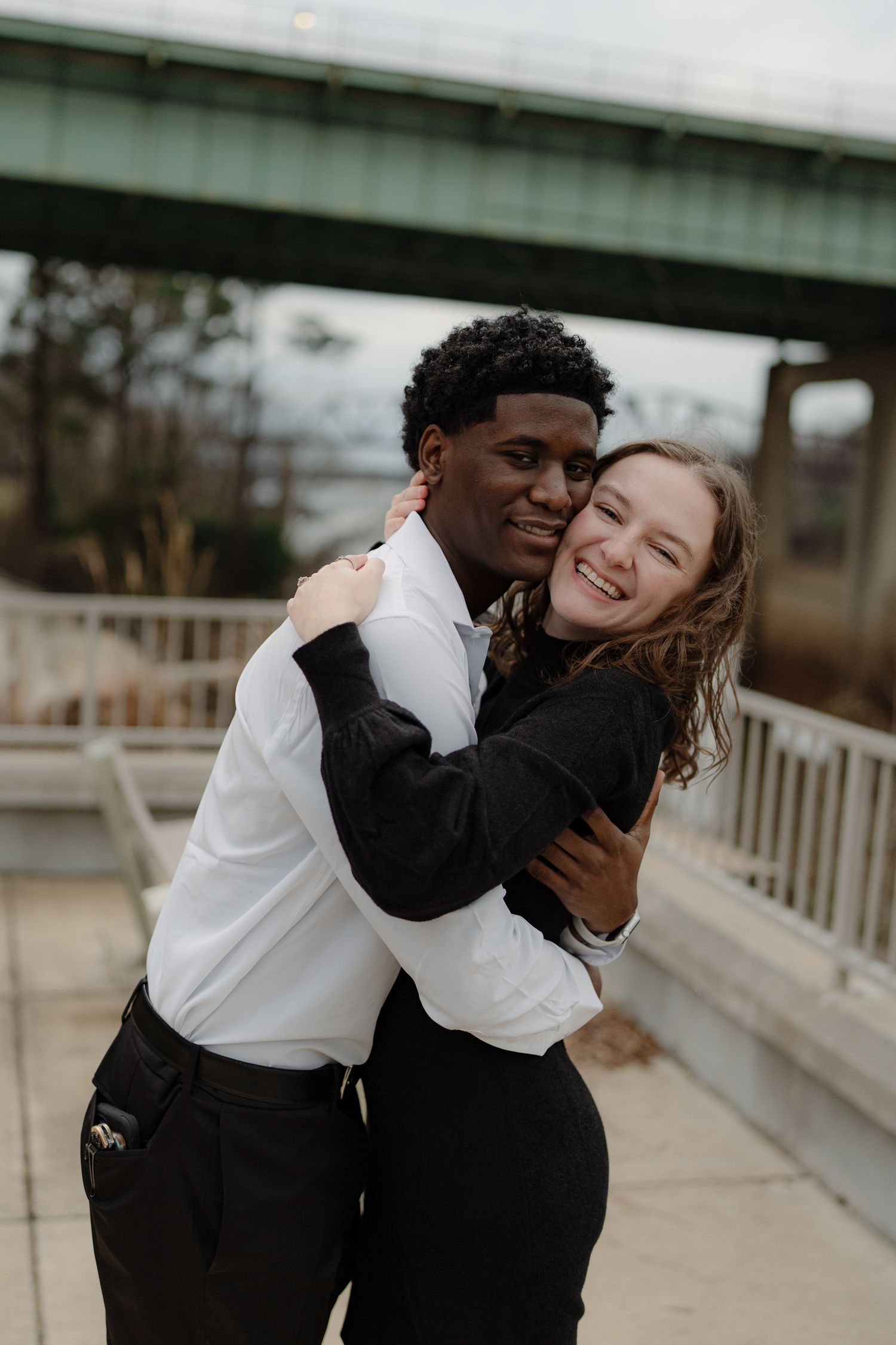 A joyful interracial couple embraces and laughs together on a bridge walkway with a green bridge behind them.