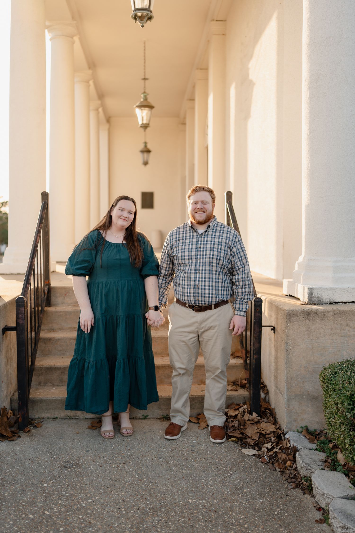 Smiling couple holding hands on steps of white columned building, woman in green dress, man in plaid.