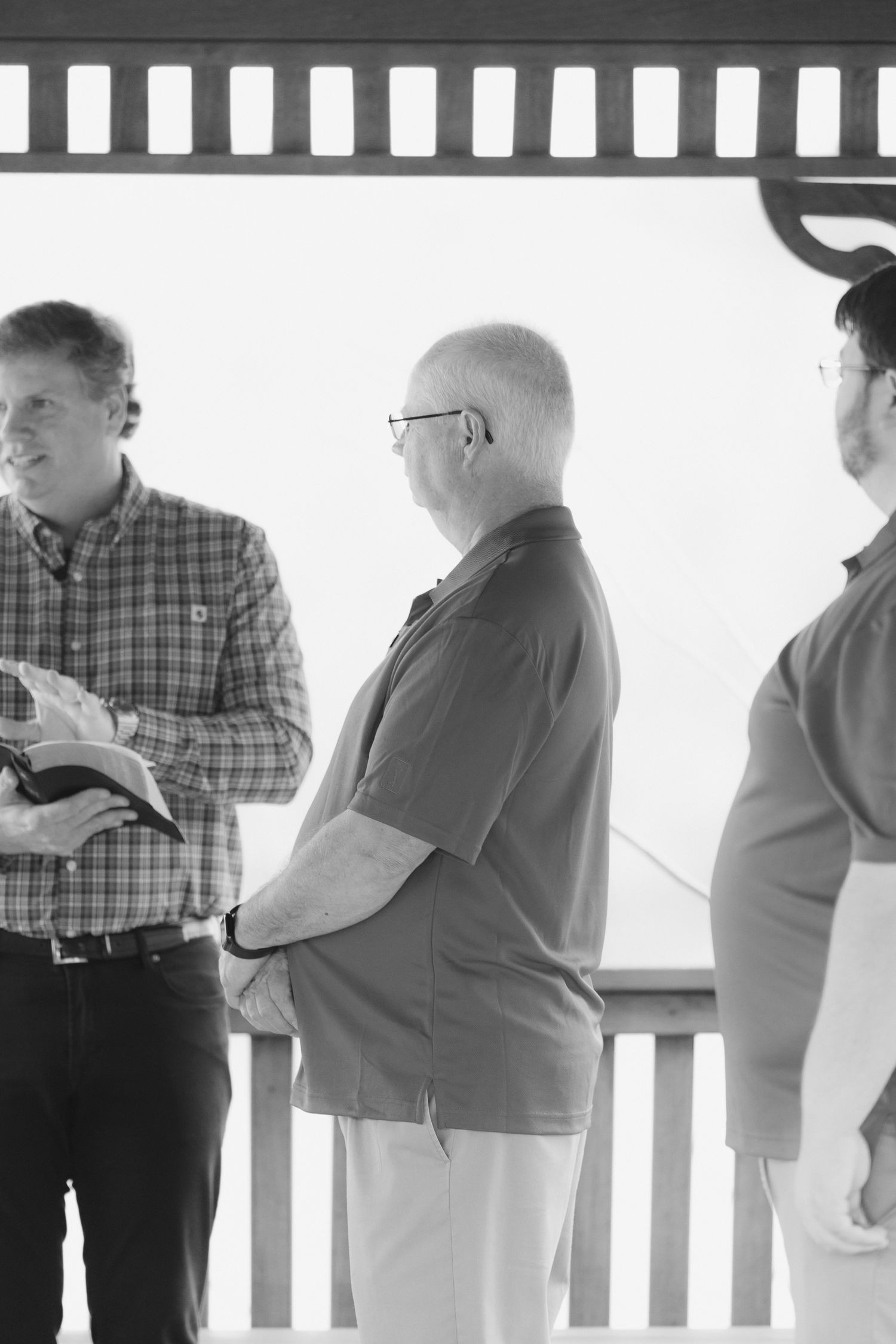 Three men stand on a porch during an outdoor ceremony, one holding a Bible.