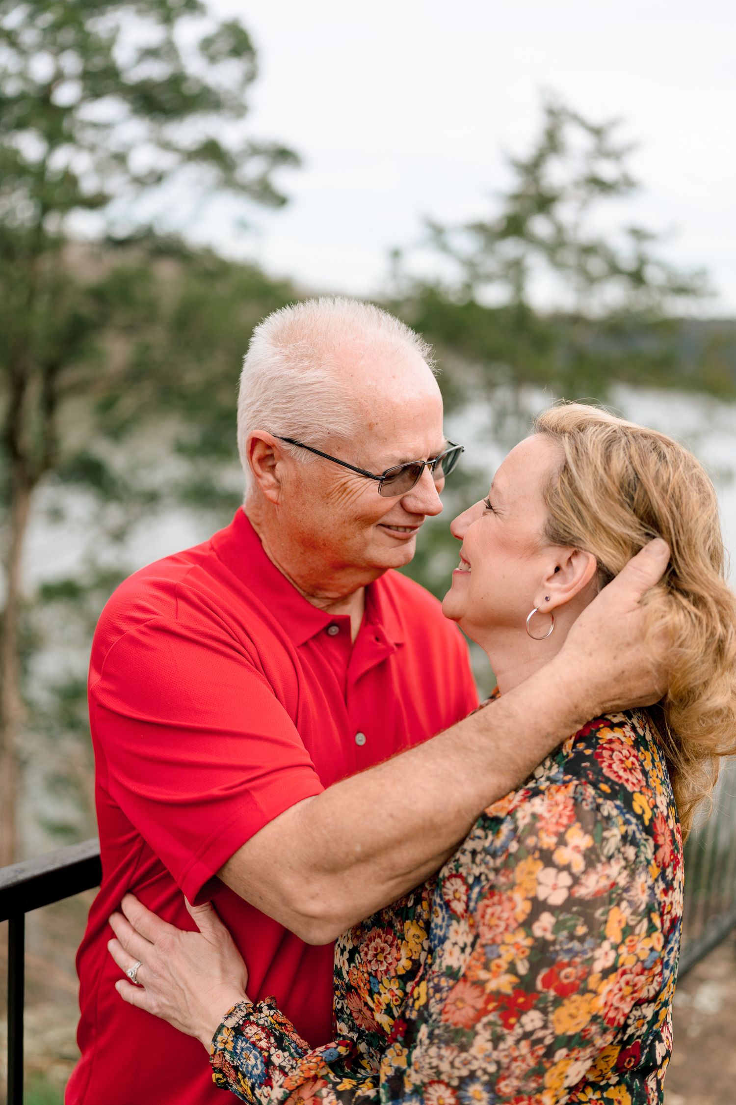 Older couple in red and floral outfits sharing a romantic moment outdoors near a lake.
