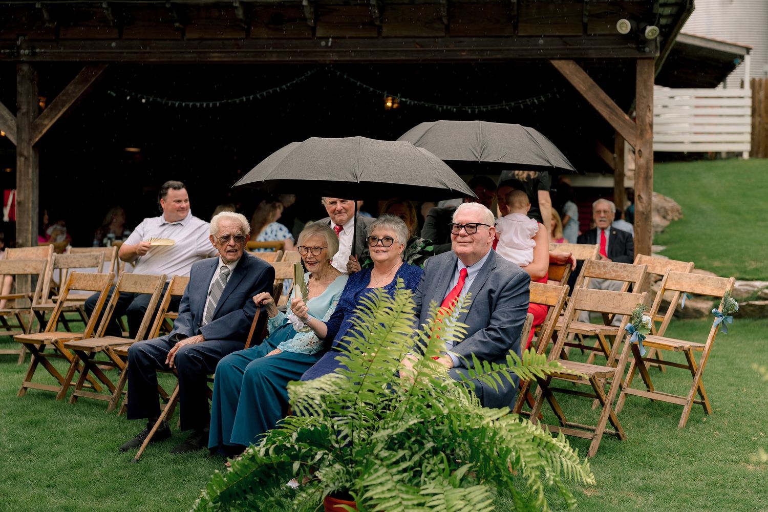 Wedding guests seated on wooden chairs outdoors, some holding umbrellas, with ferns decorating the ceremony aisle.