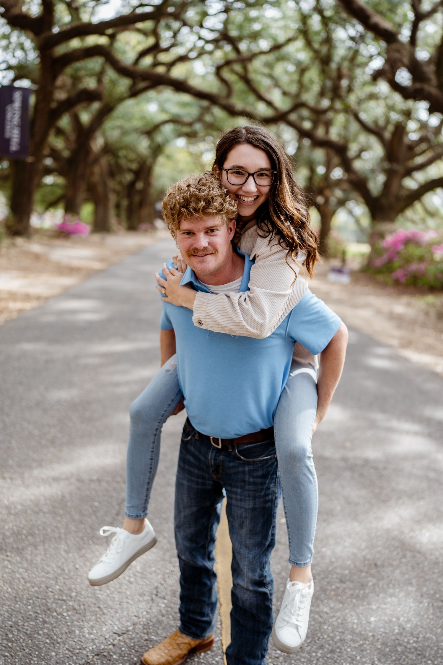 Playful couple engages in piggyback ride along tree-lined path with blue shirt and floral dress.