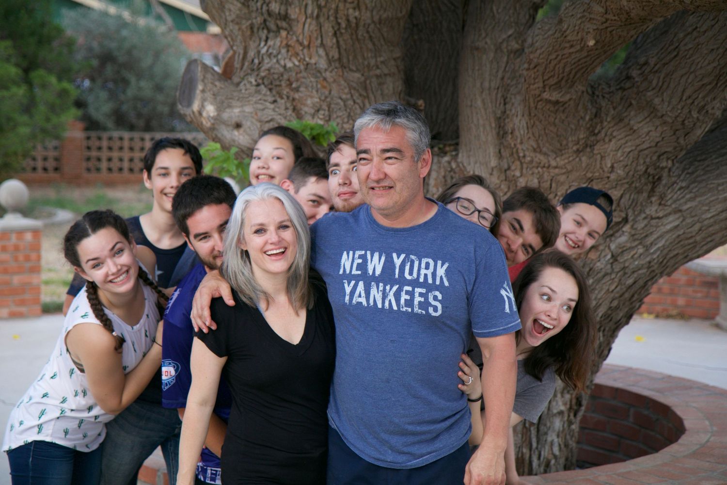 A group poses playfully around a large tree trunk, with someone wearing a New York Yankees shirt at the center.