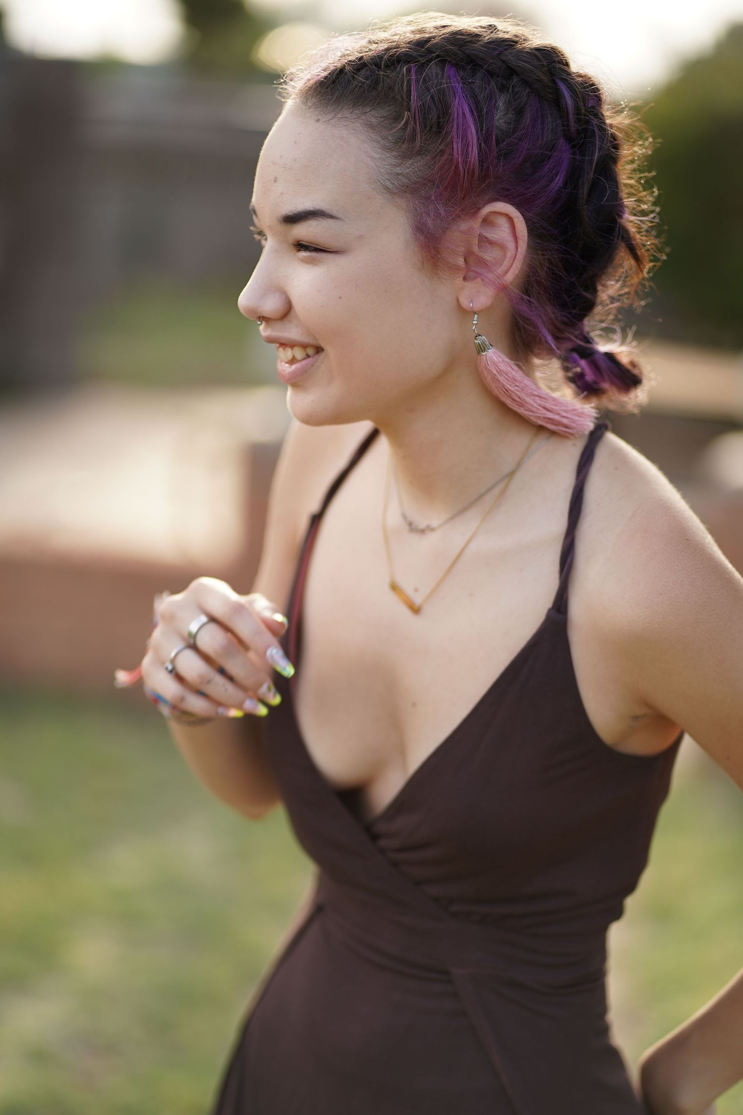 Person with pink hair wearing a black dress smiles while wearing layered necklaces outdoors.