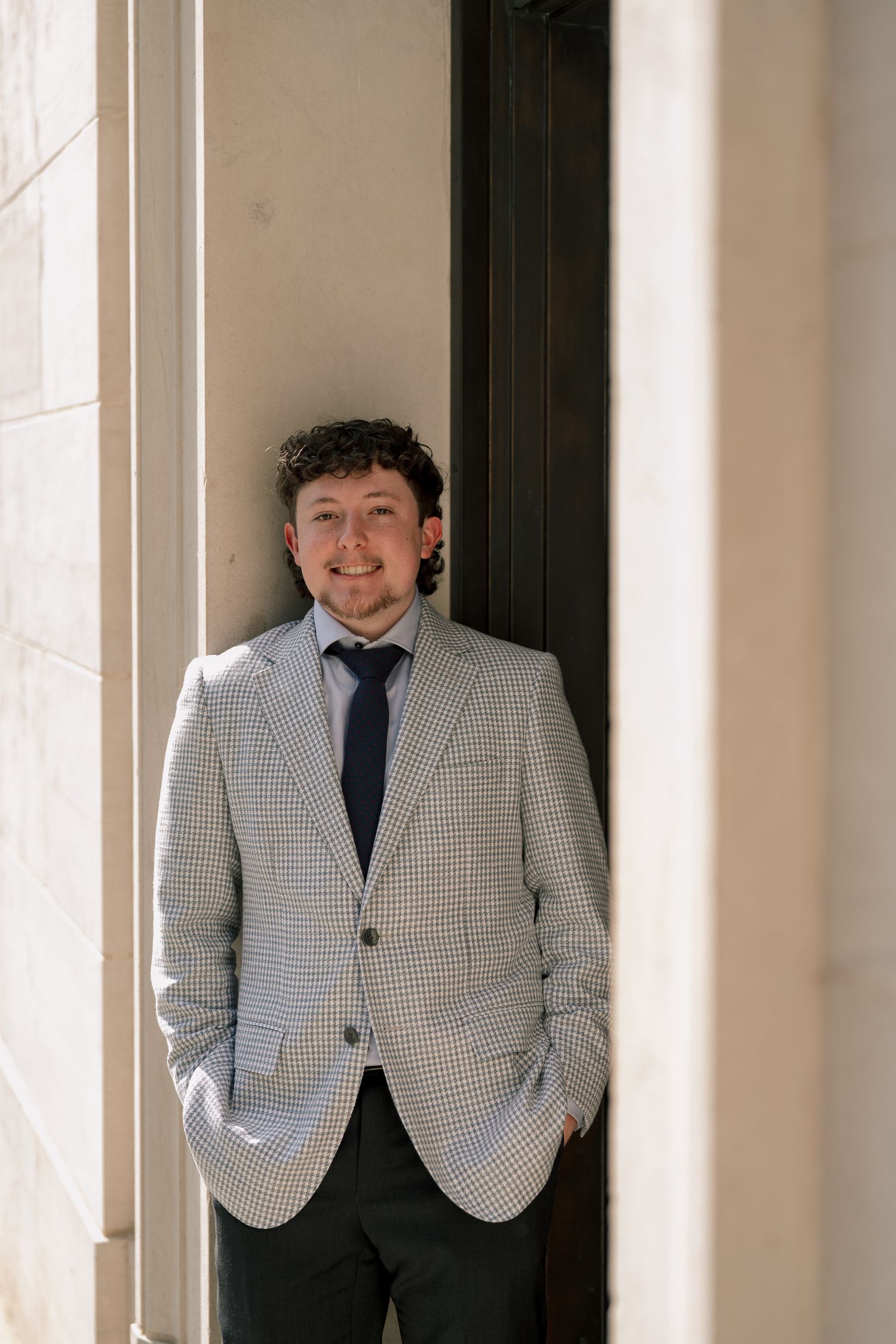 Young man in checkered blazer and tie smiling while leaning against a column in a formal outdoor setting.