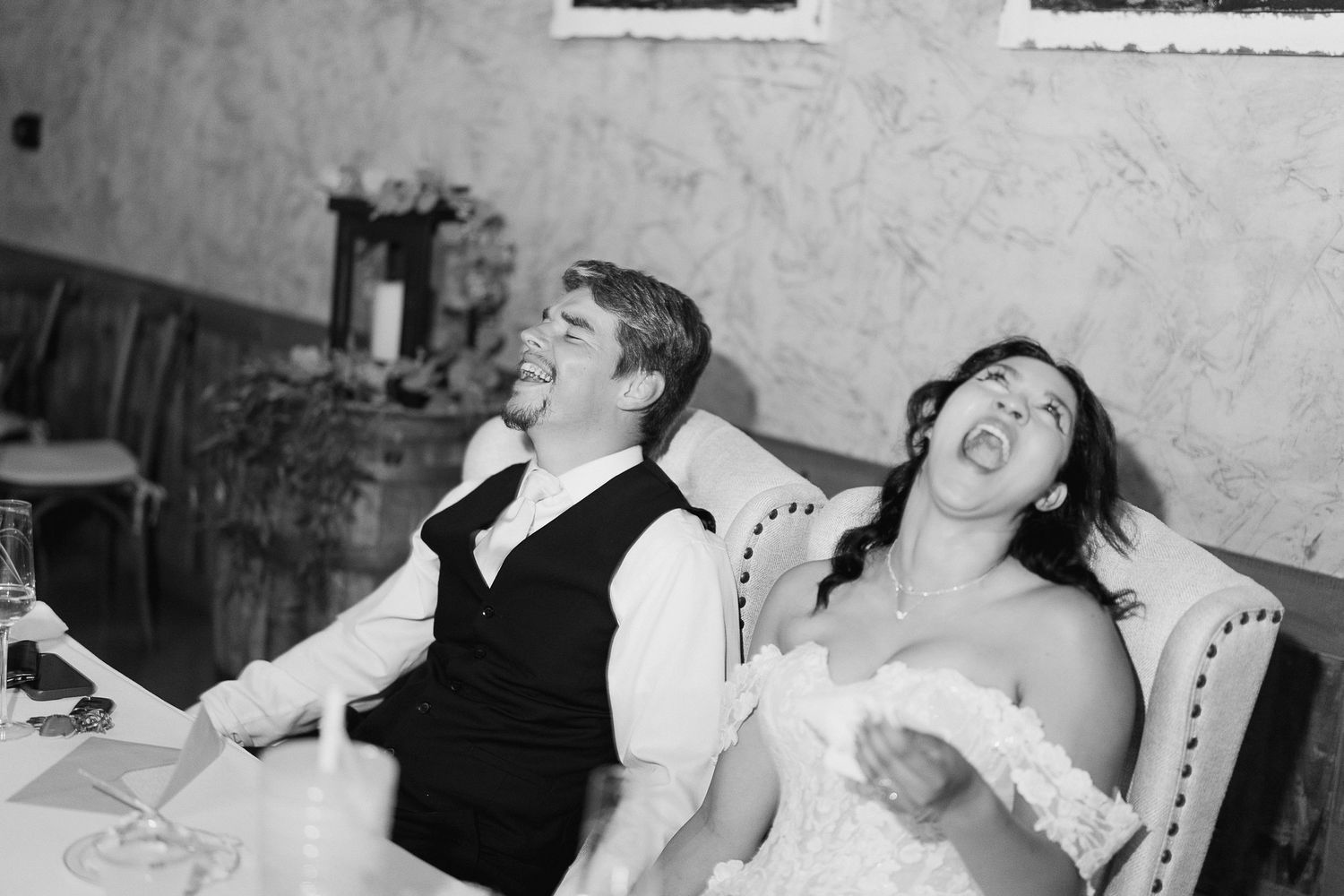 Bride and groom laugh joyfully together at wedding reception table in black and white photo.