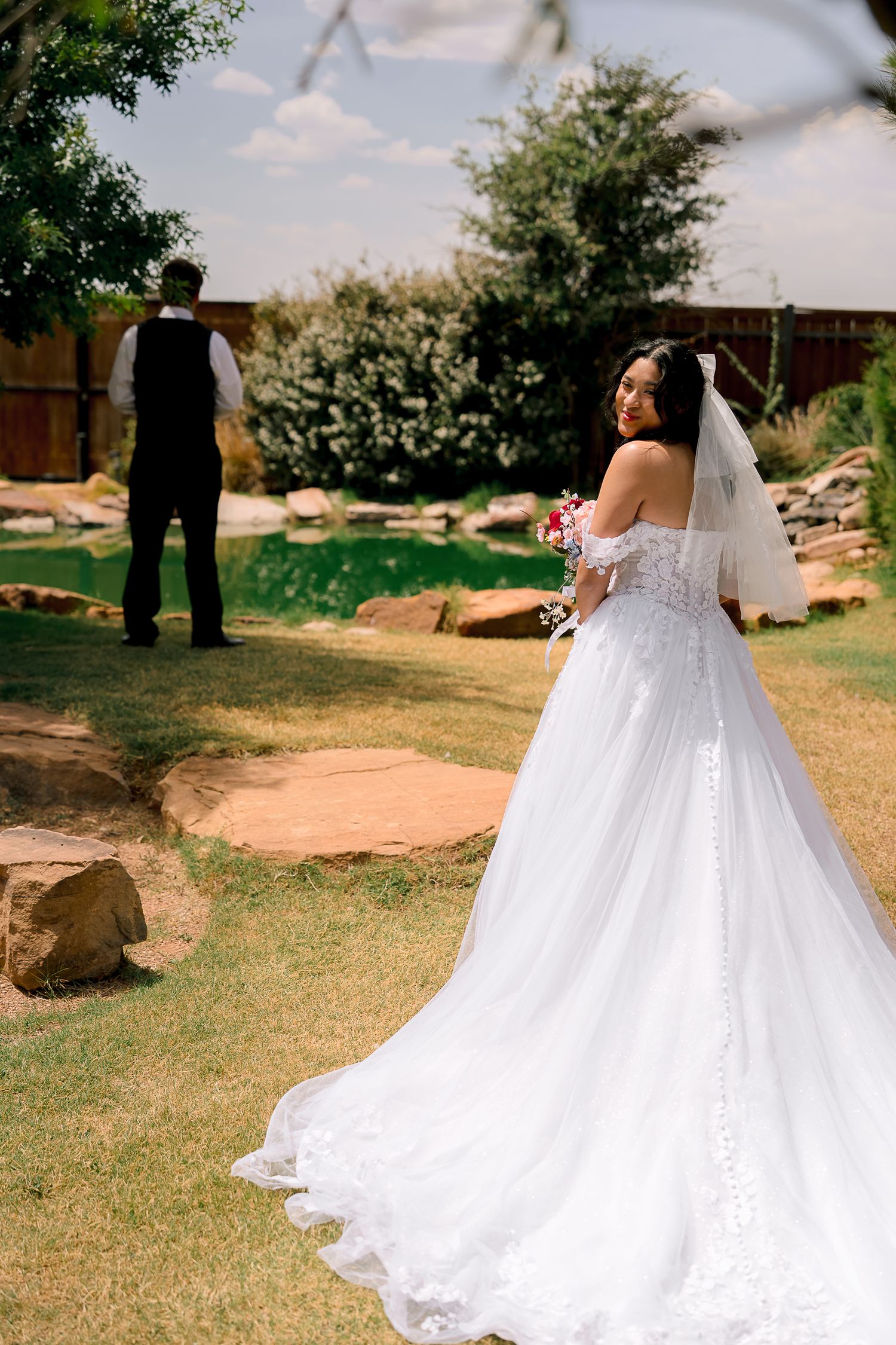 Bride in elegant white ballgown with long train holds bouquet during outdoor first look at garden wedding venue.