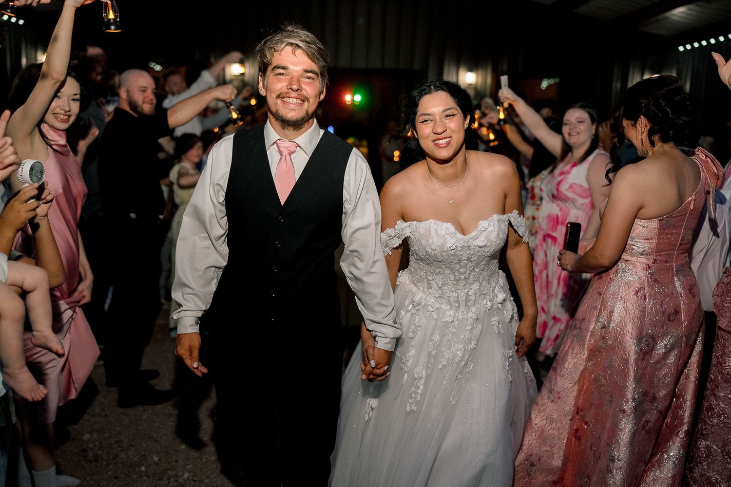 Bride and groom walk hand-in-hand through cheering guests at elegant evening wedding reception sendoff.