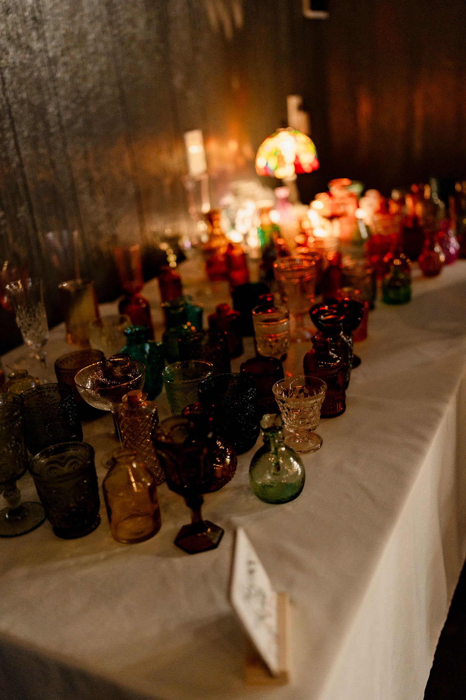 Colorful vintage glass bottles and candles line a white tablecloth creating an atmospheric glow at dusk.