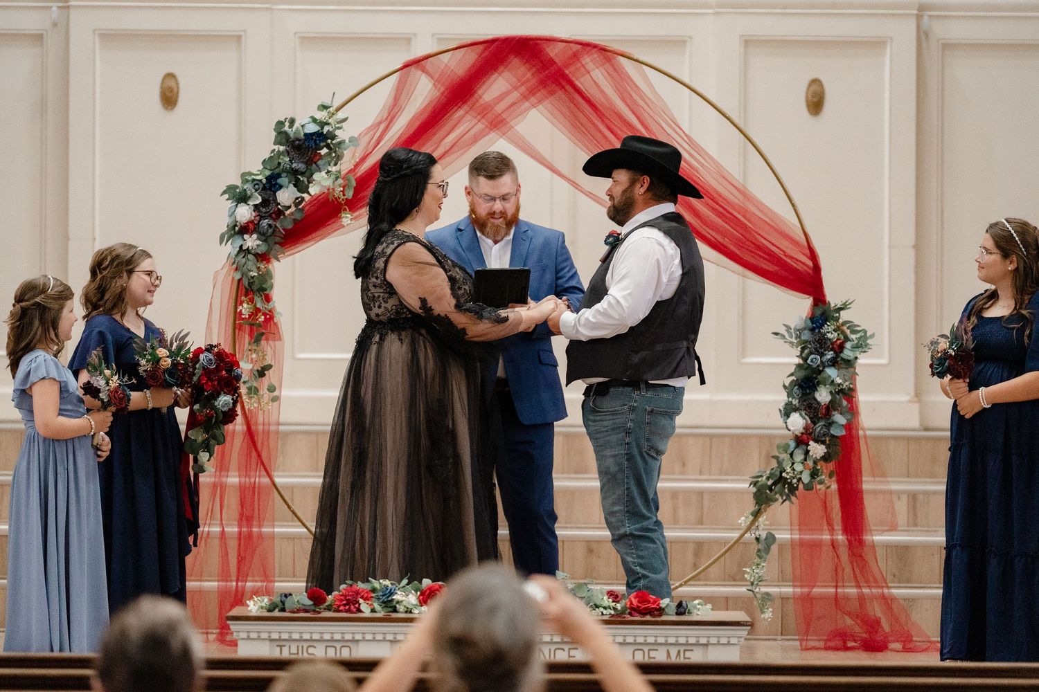Wedding ceremony guests stand in a church hall decorated with red drapes and holiday greenery.