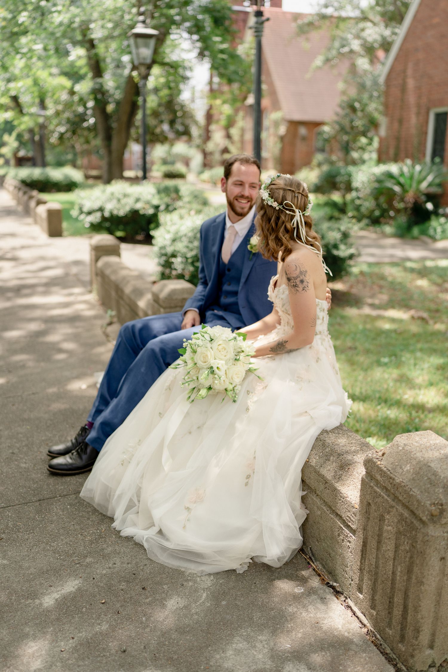 A couple in wedding attire sits together on stone steps in a garden setting during their special day.
