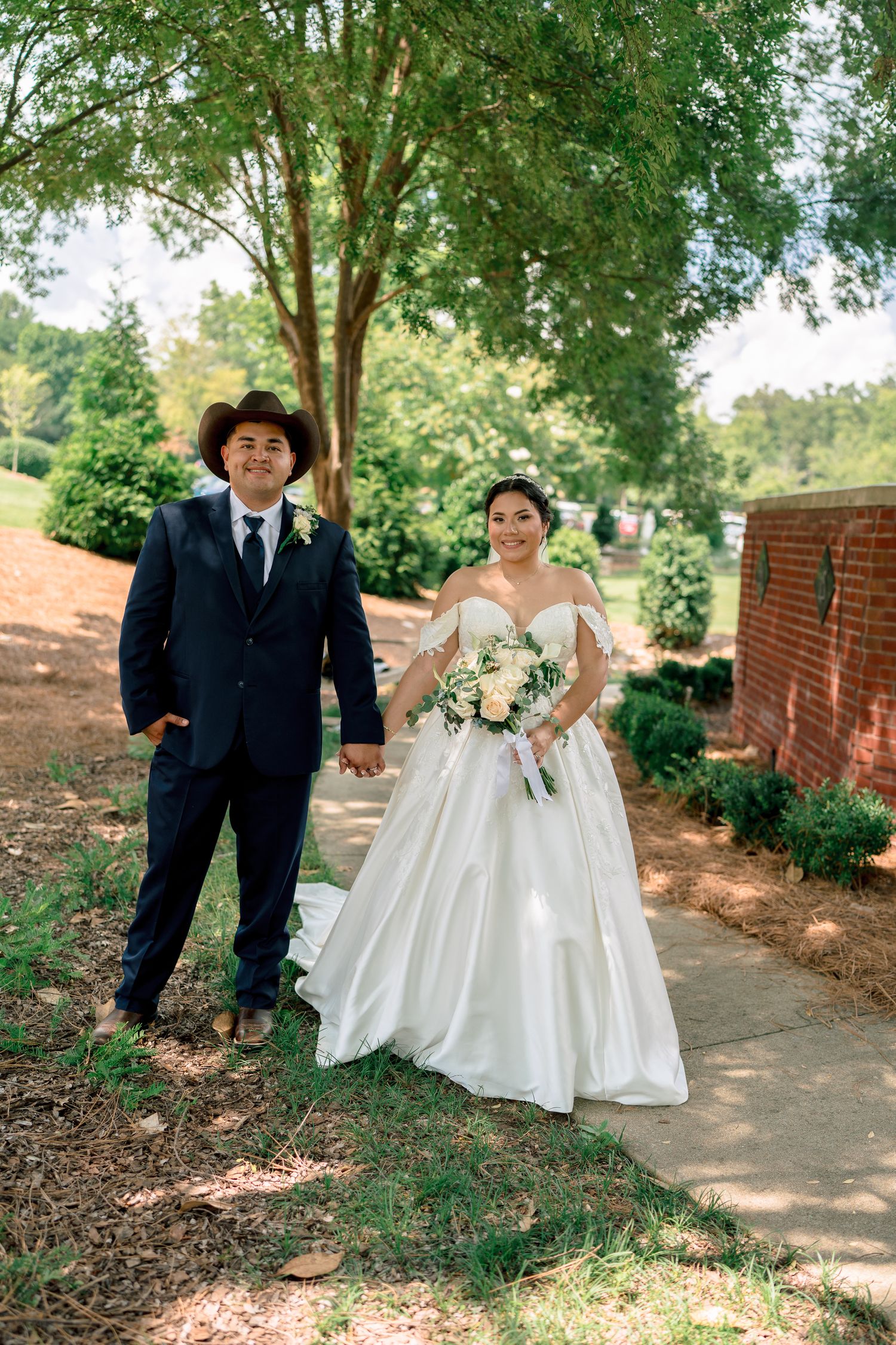 A western-themed wedding couple walking together on a garden path surrounded by trees.