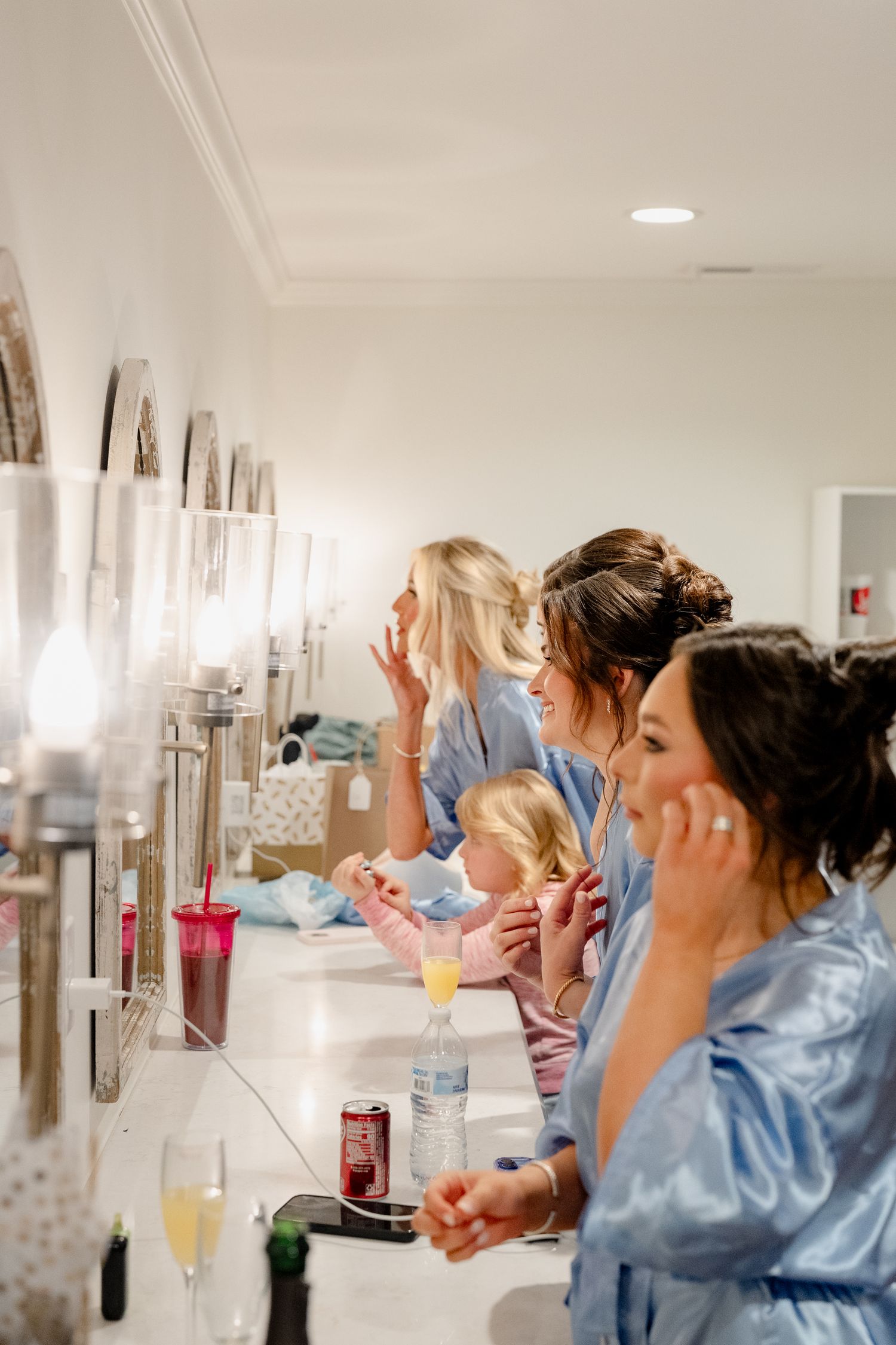 Bridesmaids in blue robes getting ready at a vanity mirror with drinks and a young child nearby.