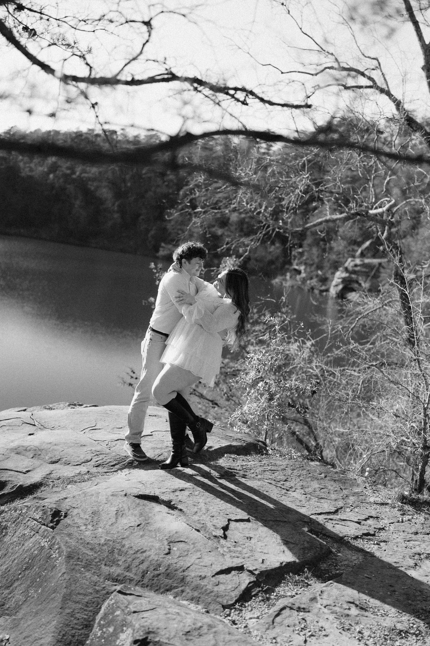 Black and white photo of couple embracing on rocky lakeside overlook amid bare trees.