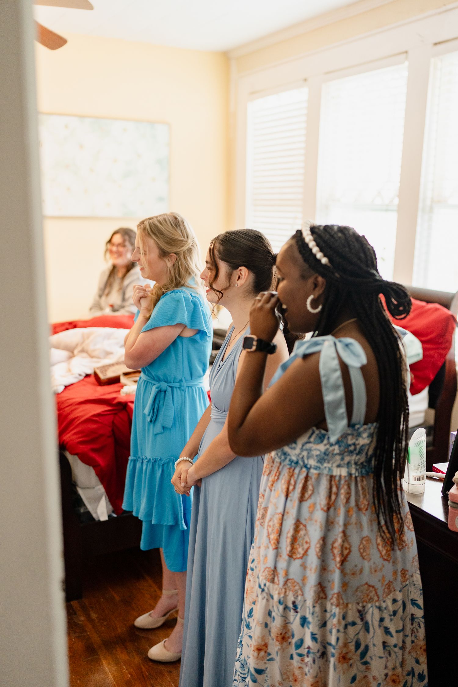 Bridesmaids in flowing blue dresses gather in a sunlit room during wedding preparations.