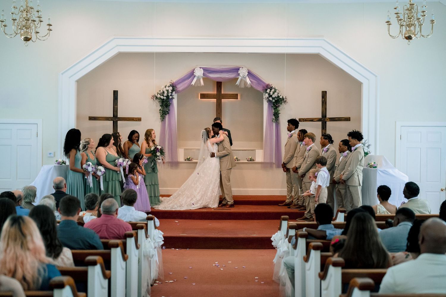 A bride and groom share their first kiss at the altar of a decorated church surrounded by wedding party members.