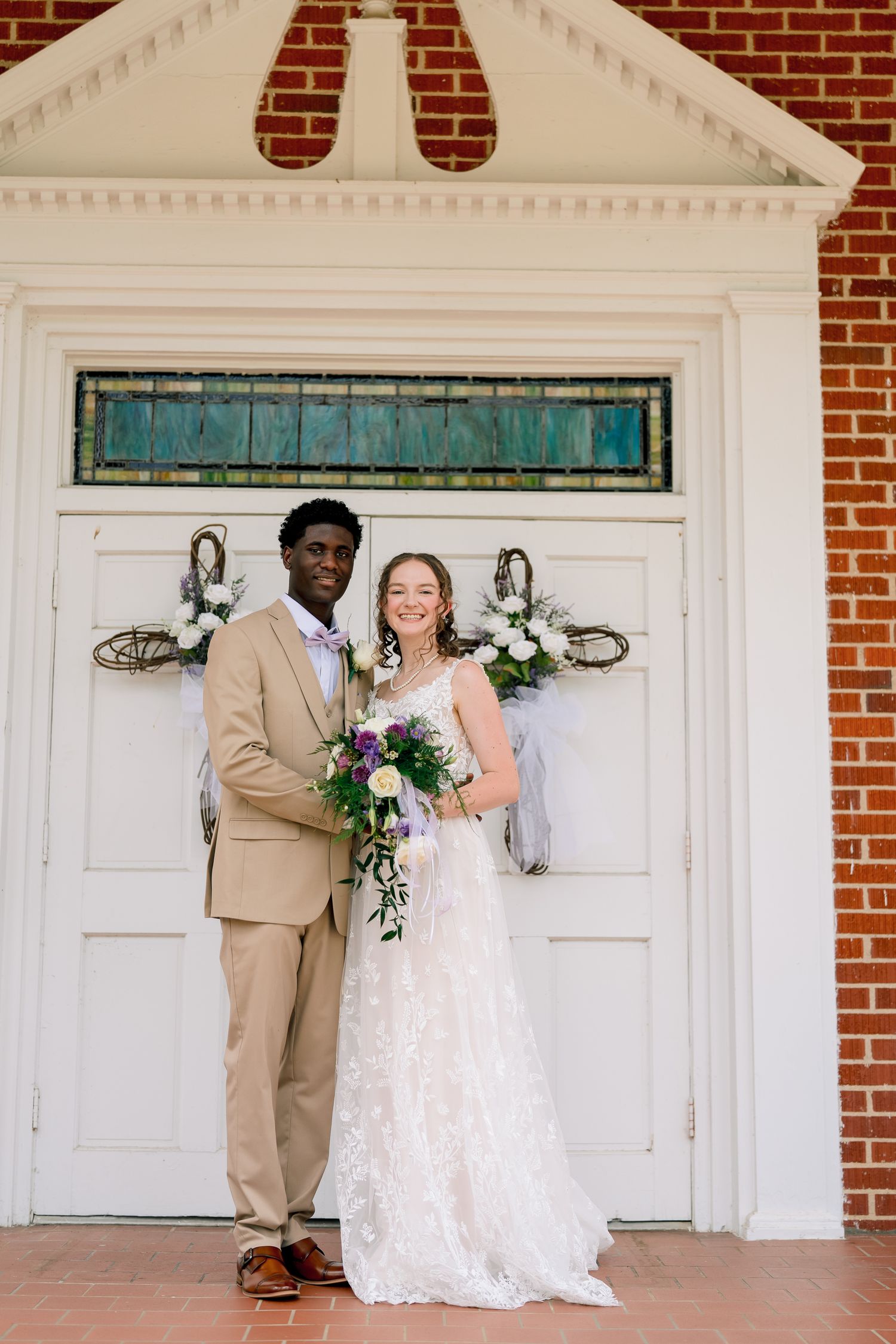Interracial couple poses in wedding attire before a white church door with floral decorations.