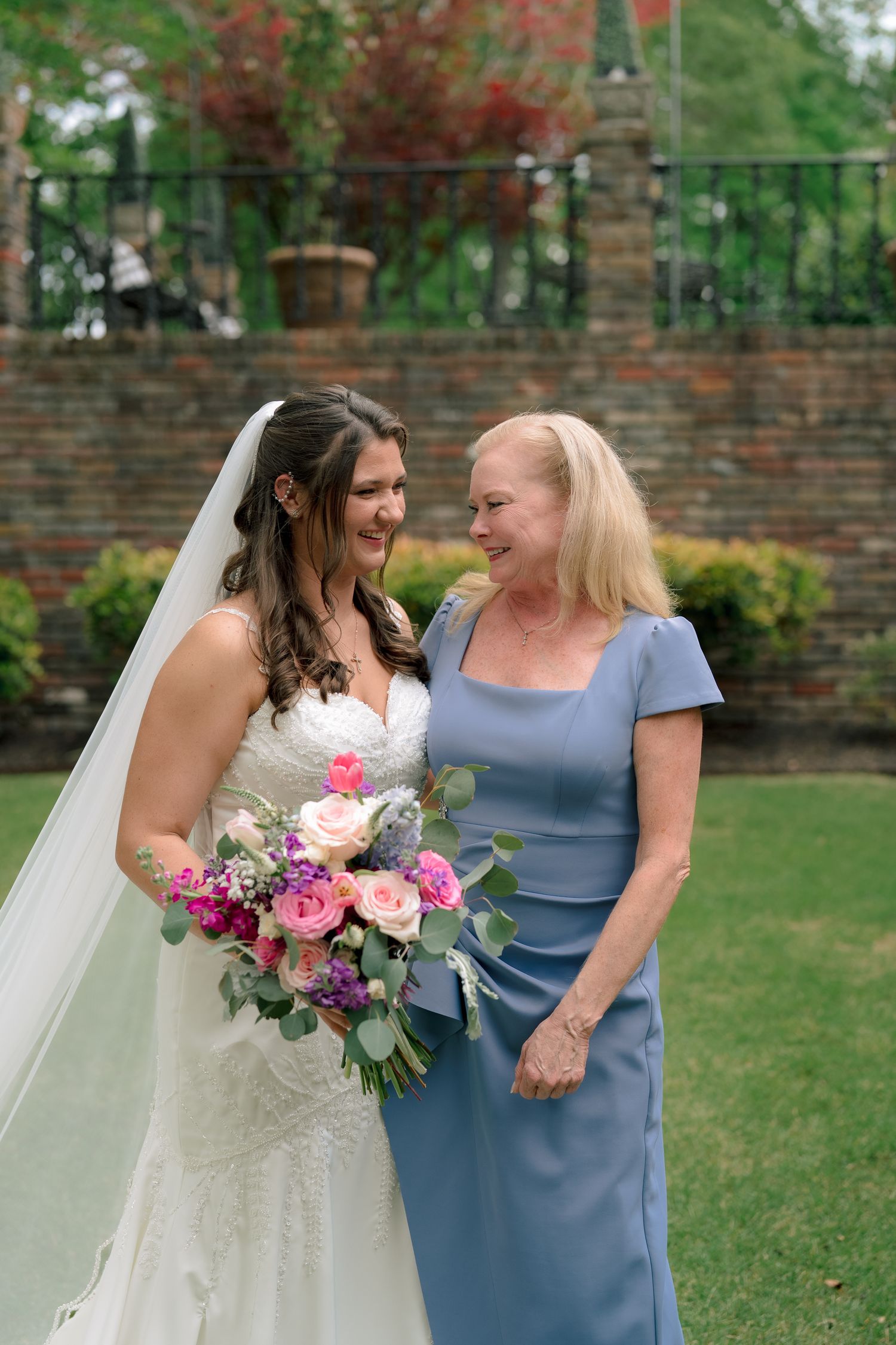 Bride in white lace gown laughs with older woman in blue dress while holding colorful bouquet.
