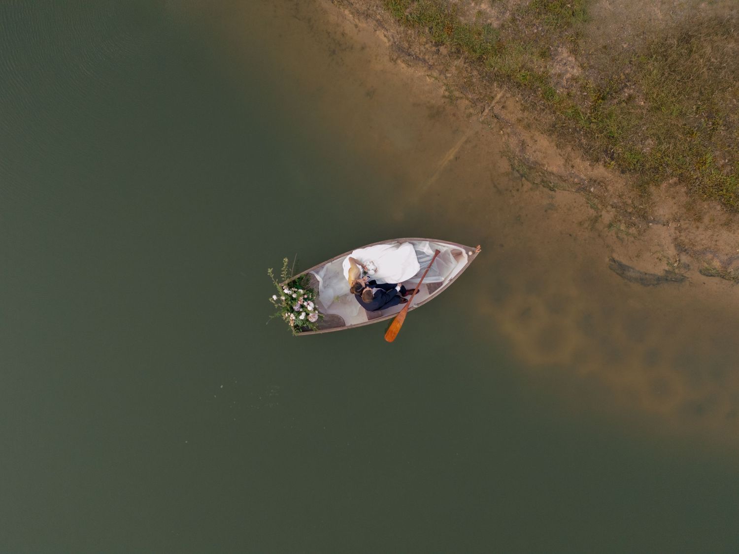 Aerial view of a person in a small wooden boat near a shallow shoreline with green vegetation.