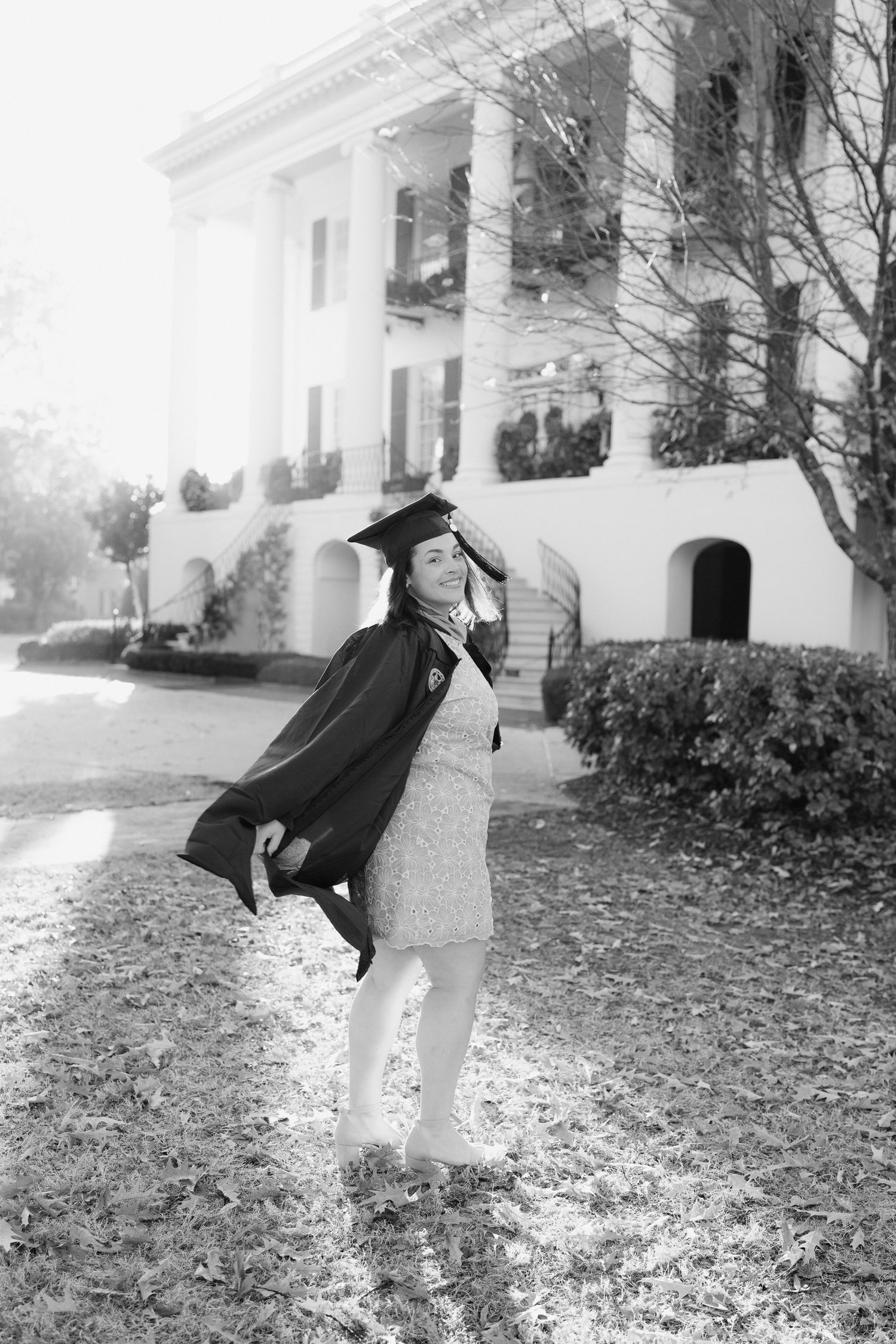 A black and white photo series shows a person in graduation attire playfully posing on a leaf-covered lawn near a building.