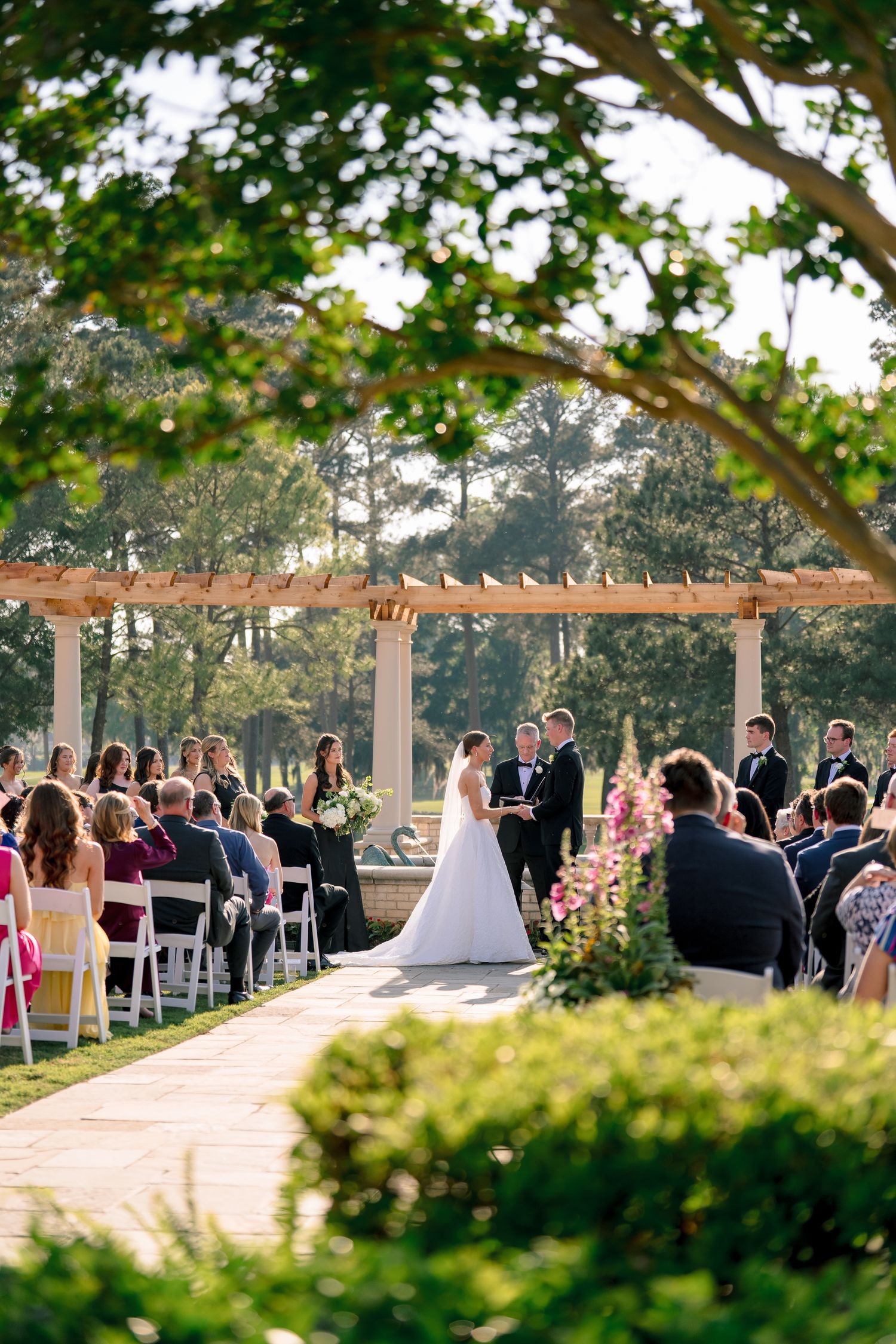 Outdoor wedding ceremony under a wooden pergola with guests seated on white chairs surrounded by lush greenery.