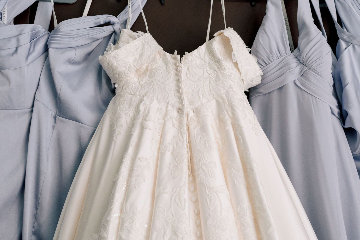 A delicate white lace flower girl dress hangs between light blue bridesmaid dresses on white hangers.