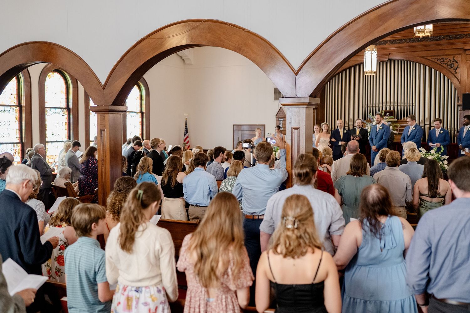 Wedding guests stand together in an elegant church interior with arched doorways and wooden organ pipes.