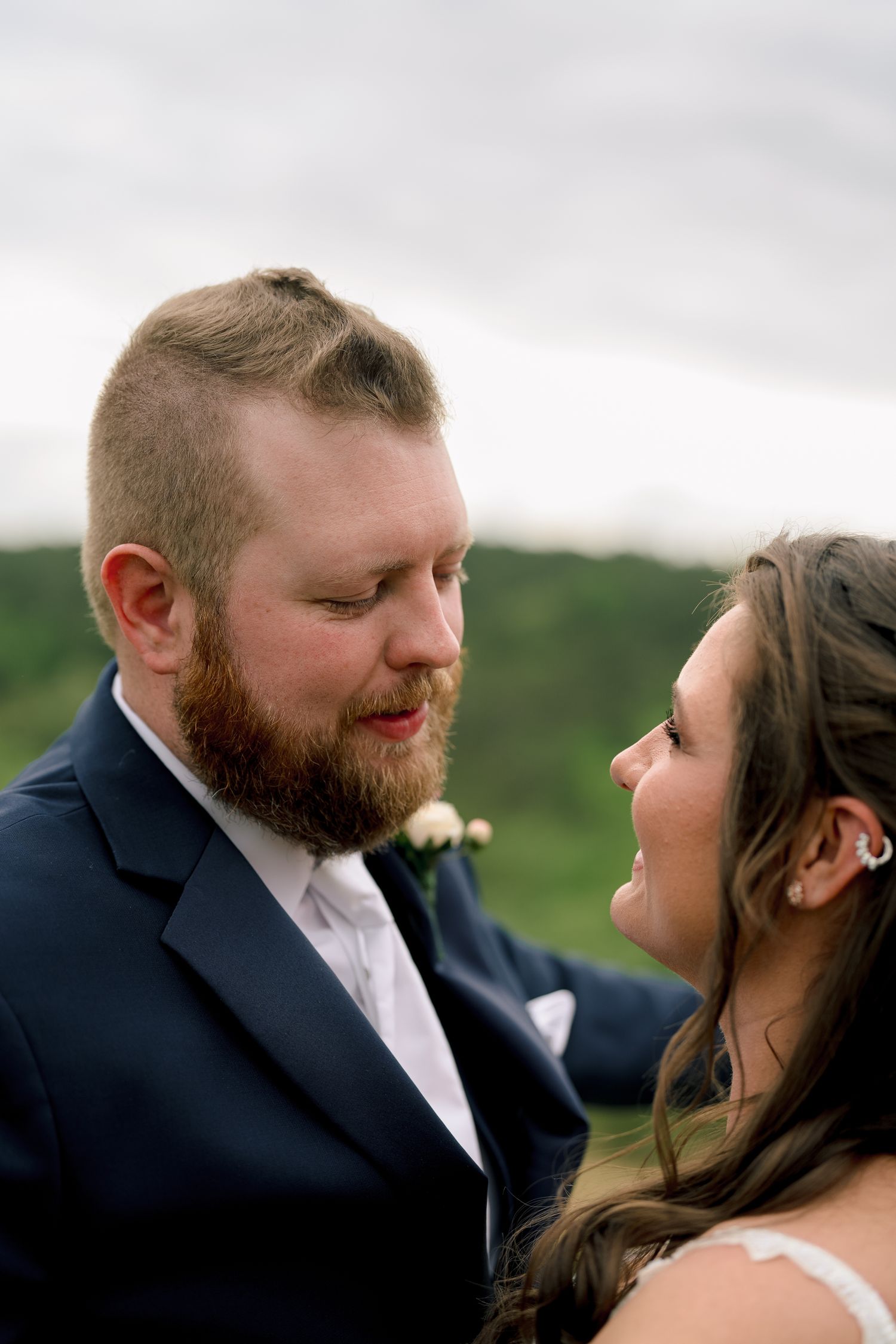 Bearded groom in navy suit gazes lovingly at smiling bride during outdoor wedding portrait.