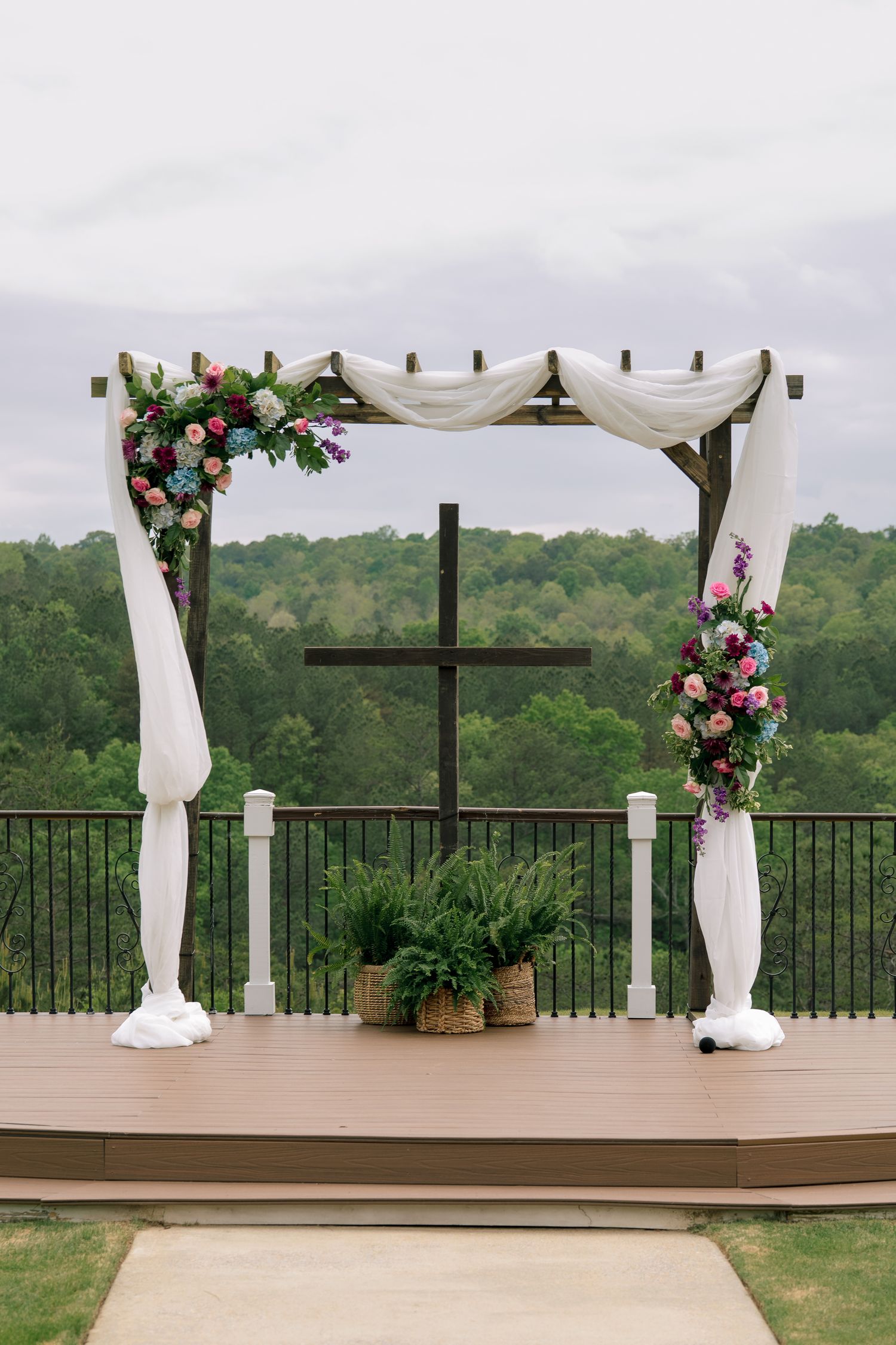 Elegant outdoor wedding arch with white draping, colorful flowers, and a wooden cross on a deck overlooking lush green forest.