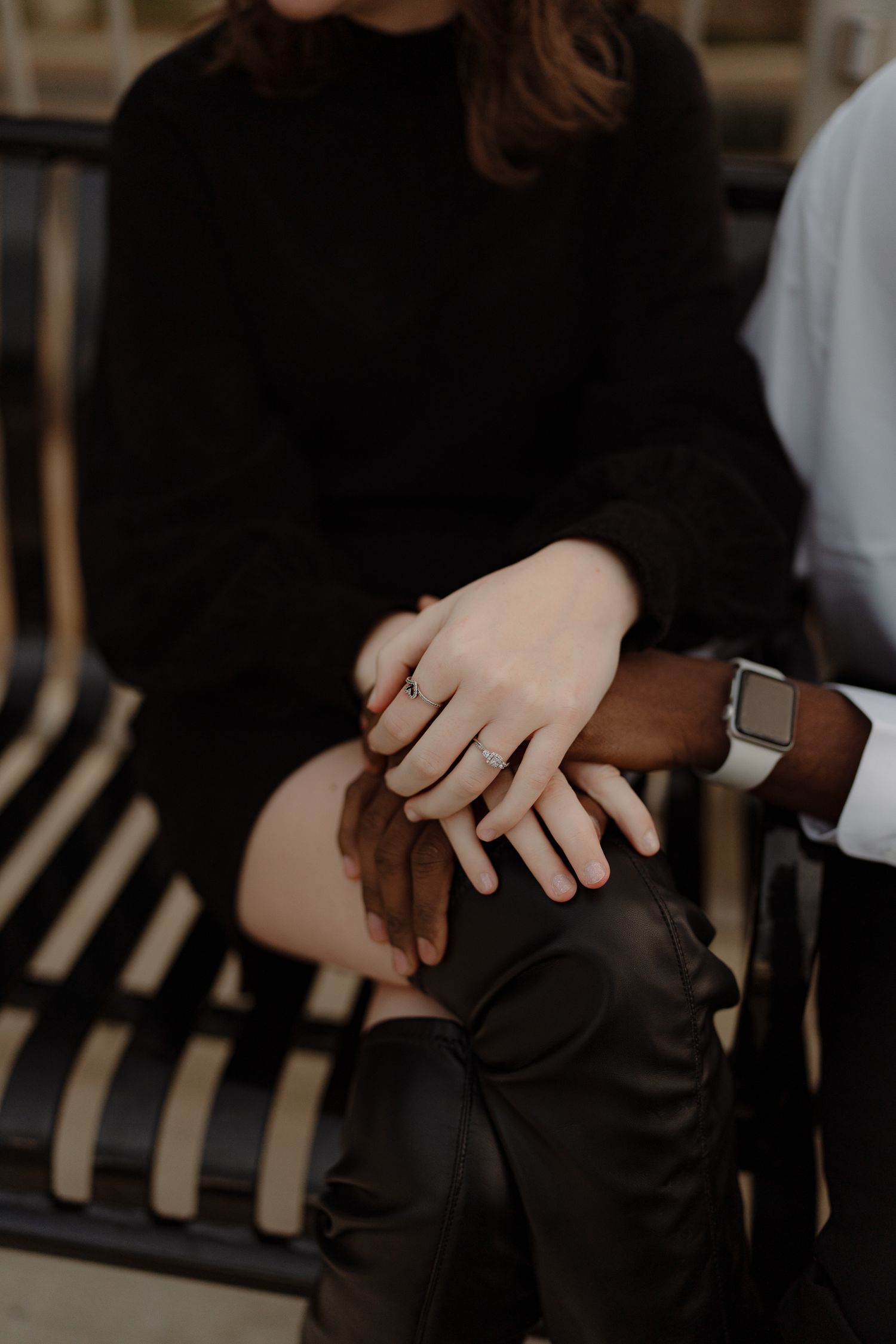 Close-up of an interracial couple holding hands, woman wearing an engagement ring, sitting together on a striped bench.