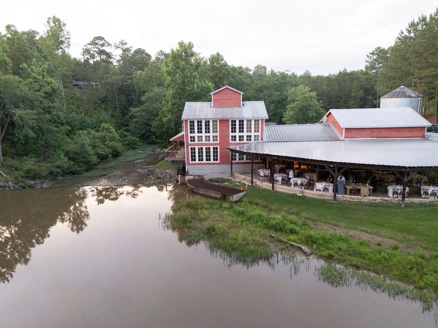 Aerial view of rustic red barn wedding venue reflecting in calm river surrounded by lush forest.