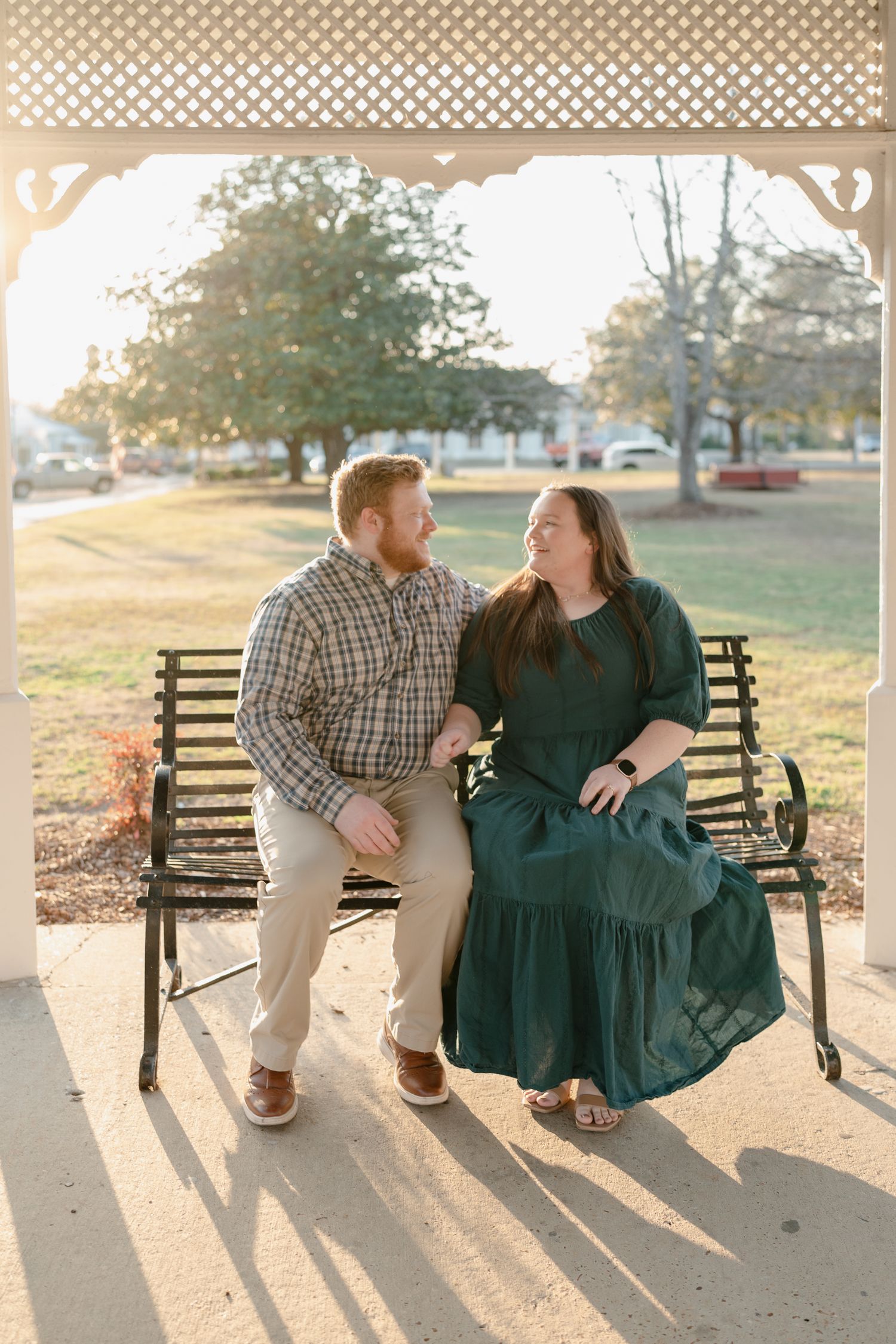 Smiling couple sitting together on a bench under a gazebo in a sunny park, sharing a loving glance.