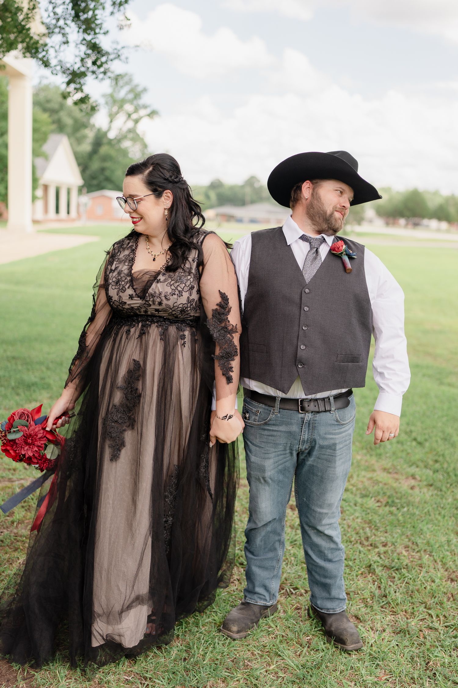 A couple in wedding attire featuring a black lace gown, cowboy hat, and red bouquet pose together on a grassy field.