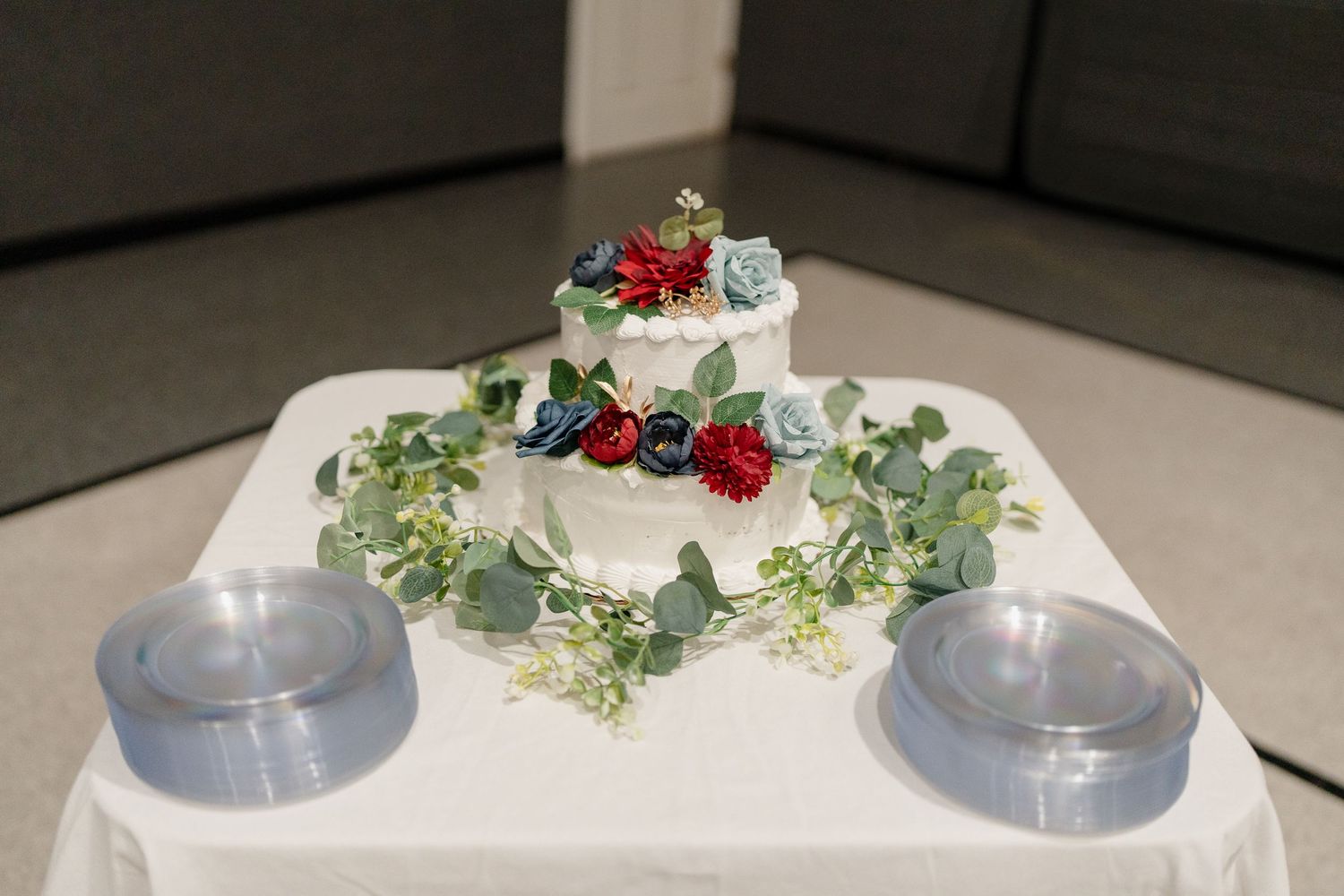 White wedding cake decorated with greenery and red floral accents on a display stand.