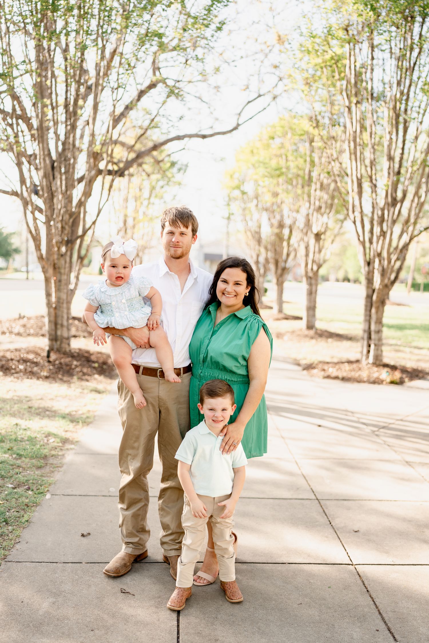 A family of four poses together on a sunlit pathway surrounded by trees.