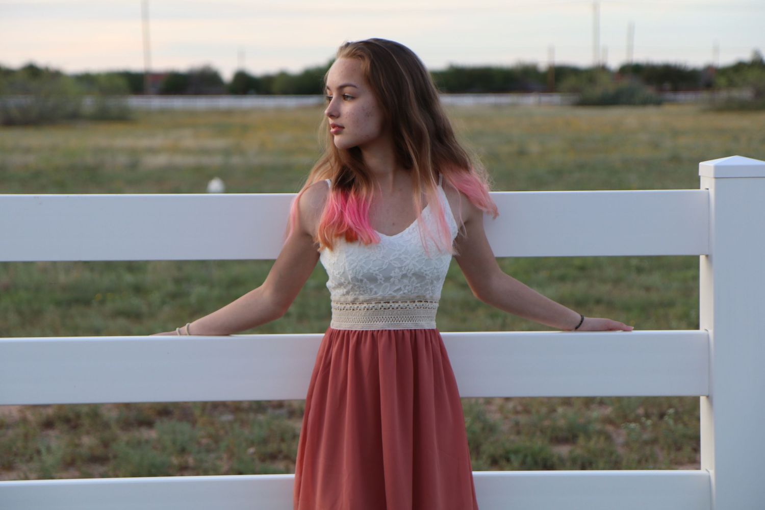 Woman in coral dress leaning against white fence during sunset.