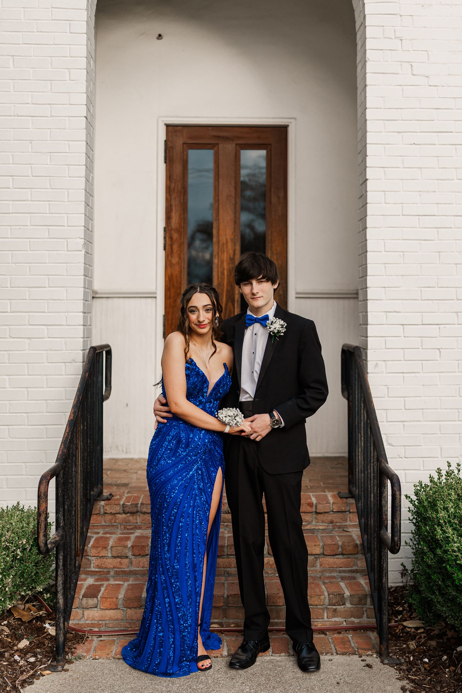 Formal couple poses in front of wooden door entrance with white brick and metal railings.