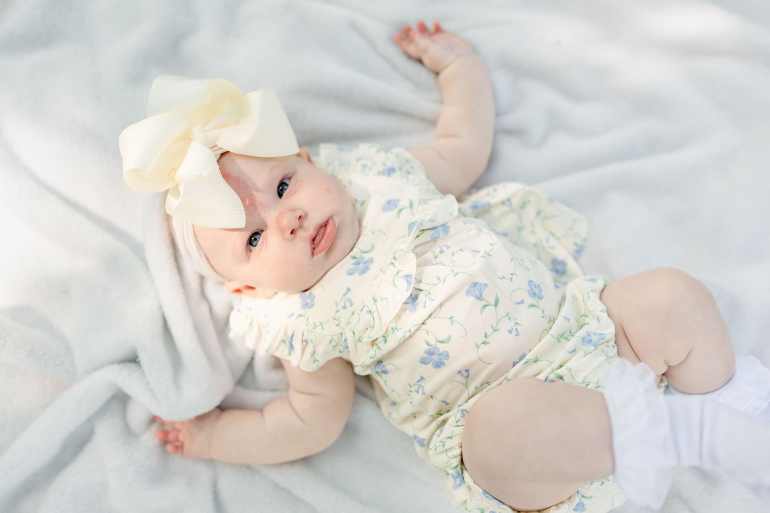 Baby in floral outfit resting on white blanket during outdoor photo session.