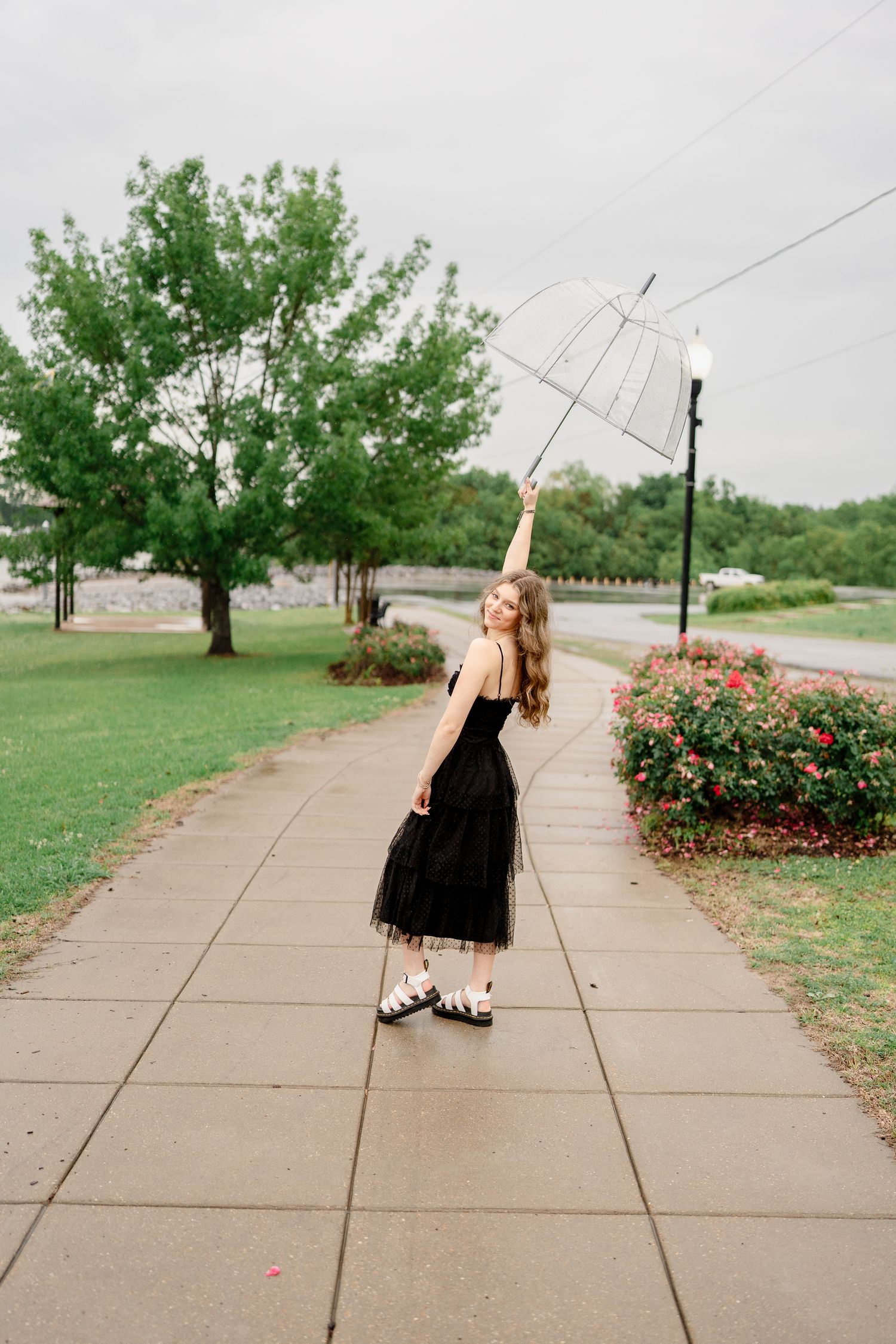 A figure in a black dress stands on a path with an umbrella, surrounded by greenery and landscaping.