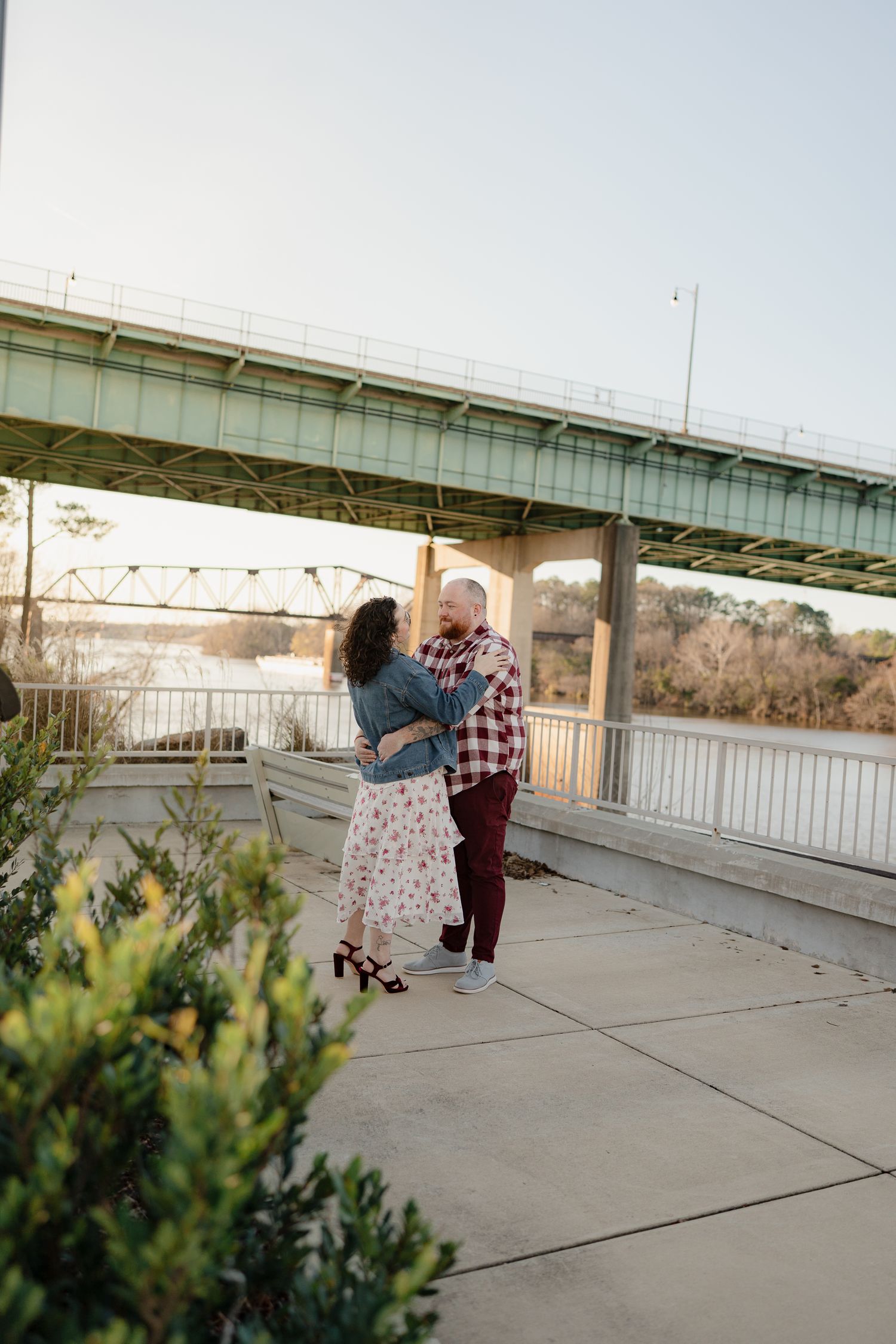 A couple embraces while standing on a concrete walkway near a bridge structure.