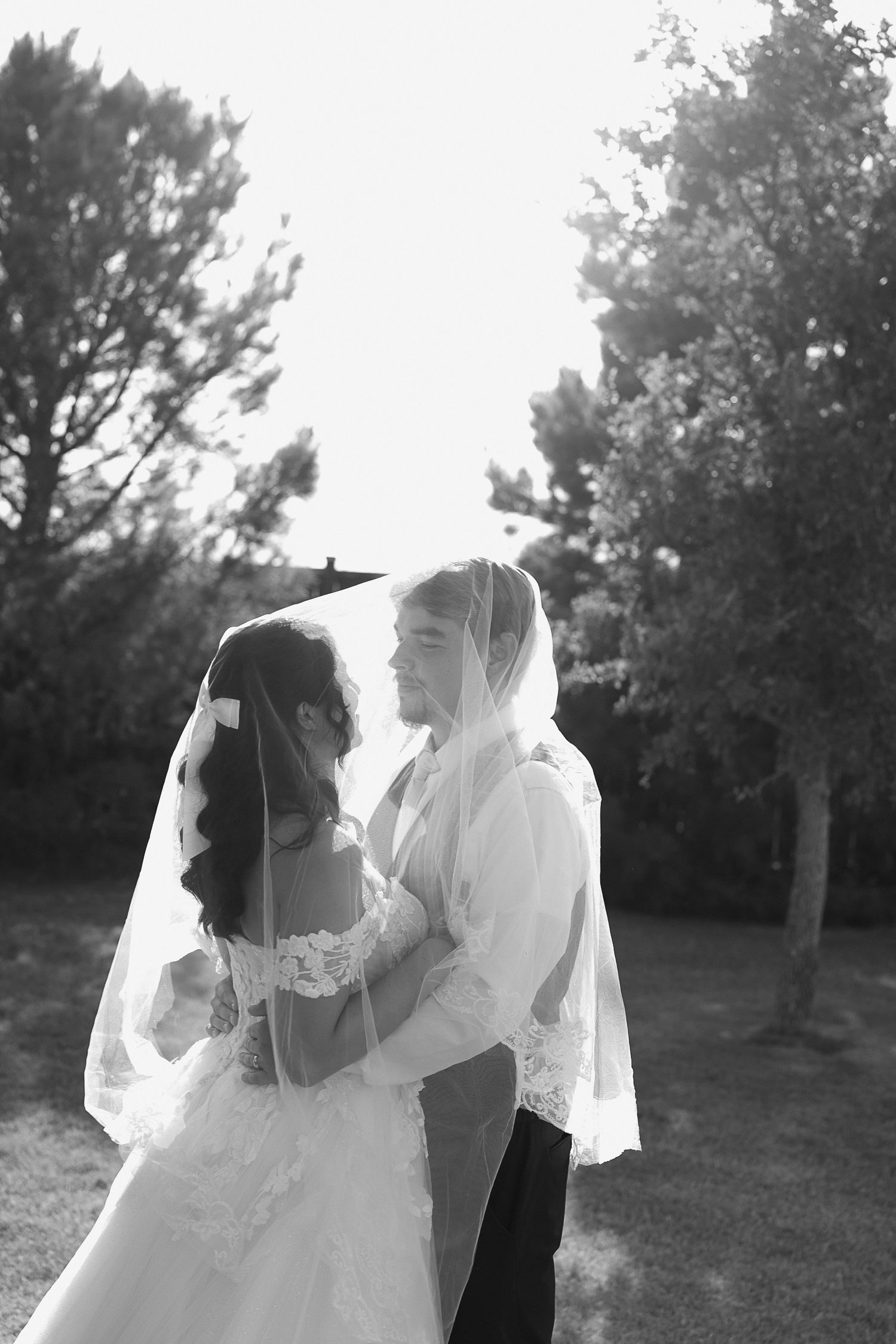 Black and white photo of bride and groom sharing intimate moment under flowing wedding veil outdoors.